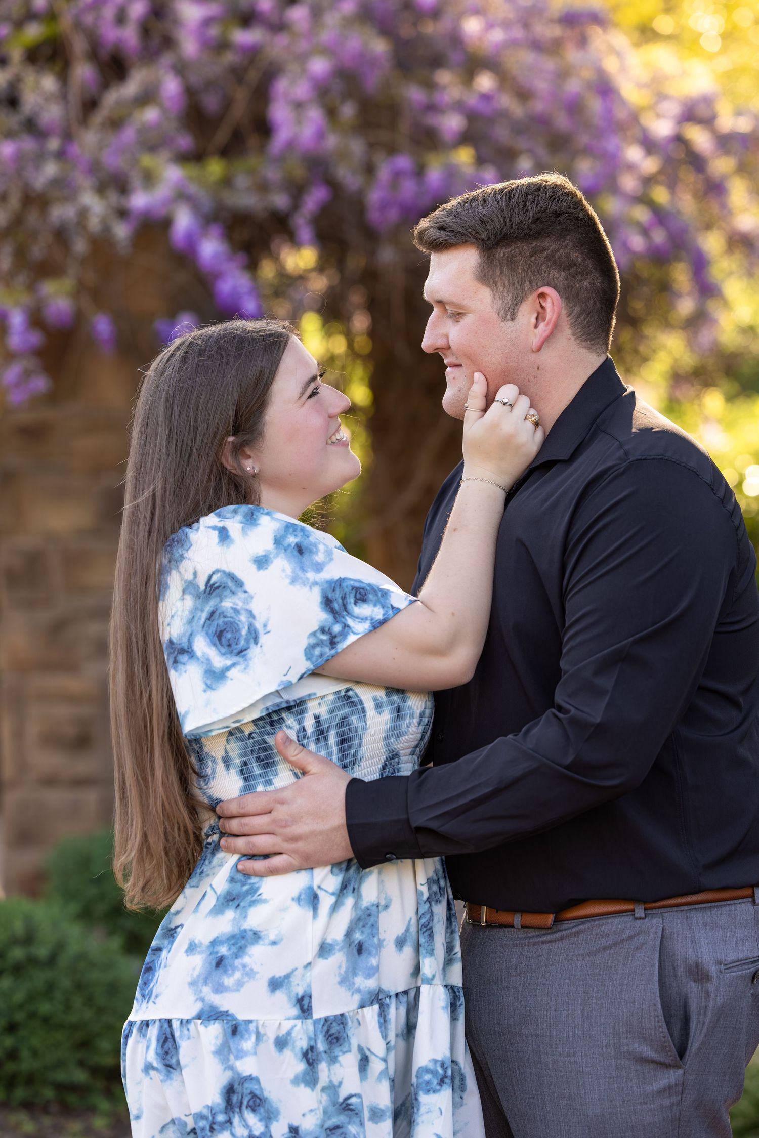 Couple embracing outdoors, woman in floral dress touching man's face, smiling, with purple flowering tree behind them.