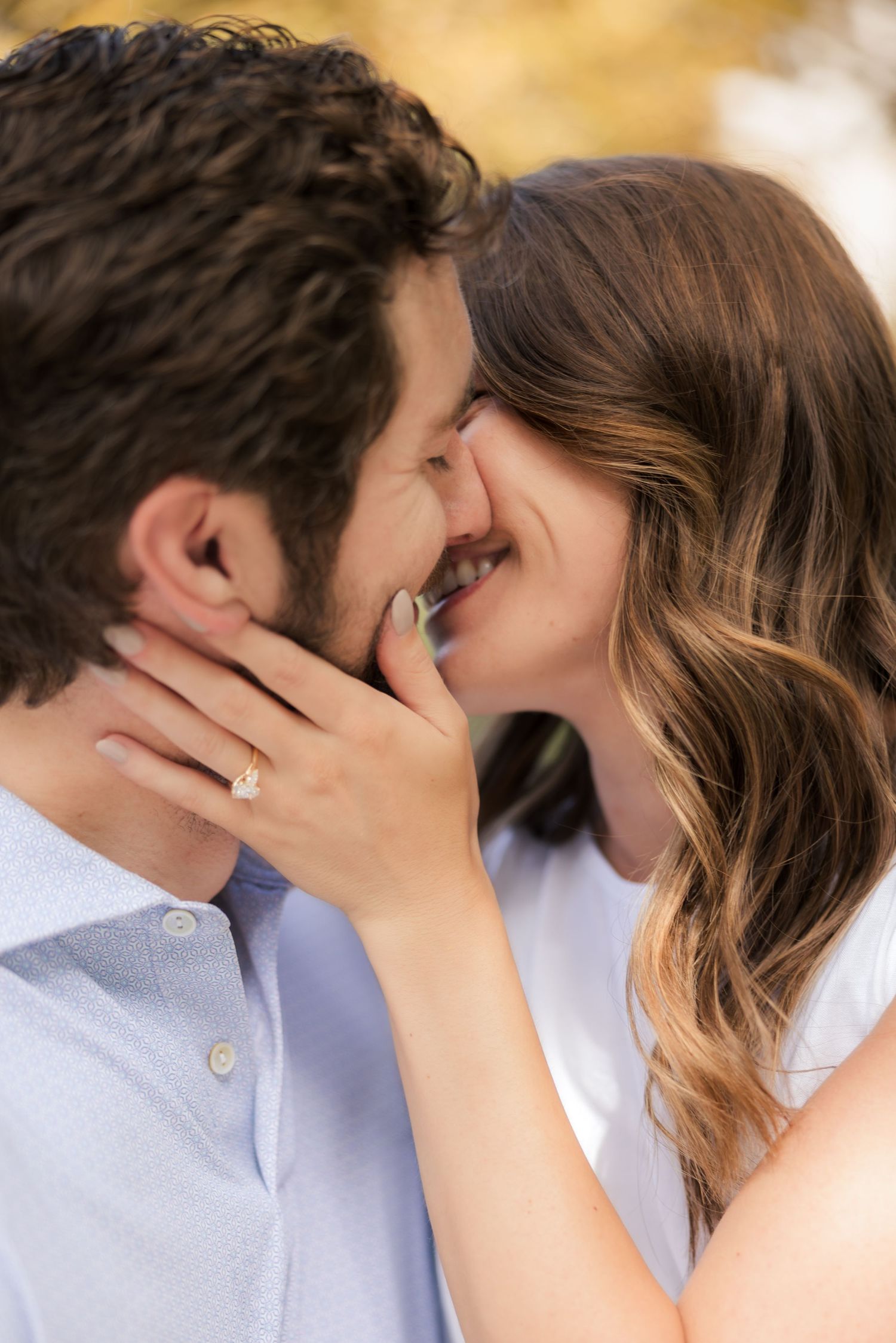 A couple shares a romantic almost-kiss moment outdoors, with the woman displaying a diamond engagement ring on her hand.
