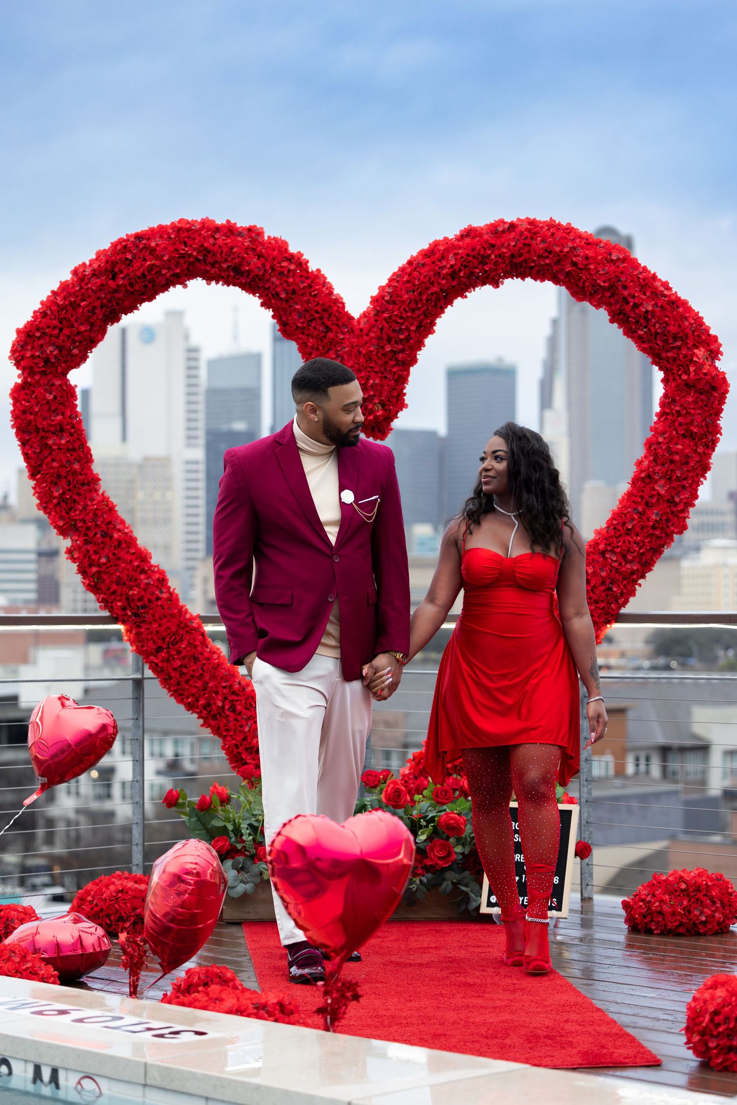 Couple holding hands in front of a large red rose heart arch with city skyline, red balloons, and Valentine's Day decor.