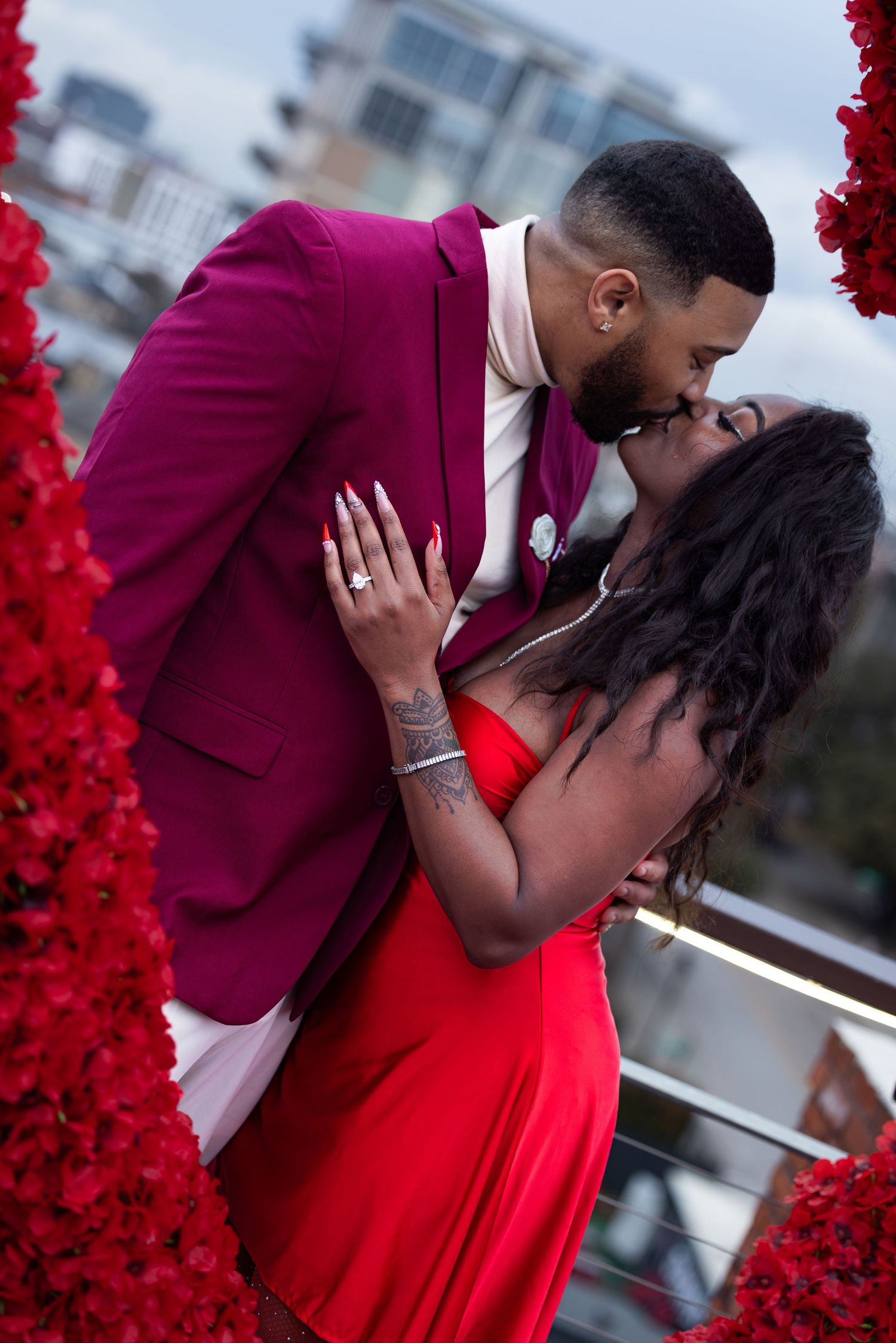 A romantic couple in burgundy and red formal attire share an intimate moment on a balcony with red floral decorations.