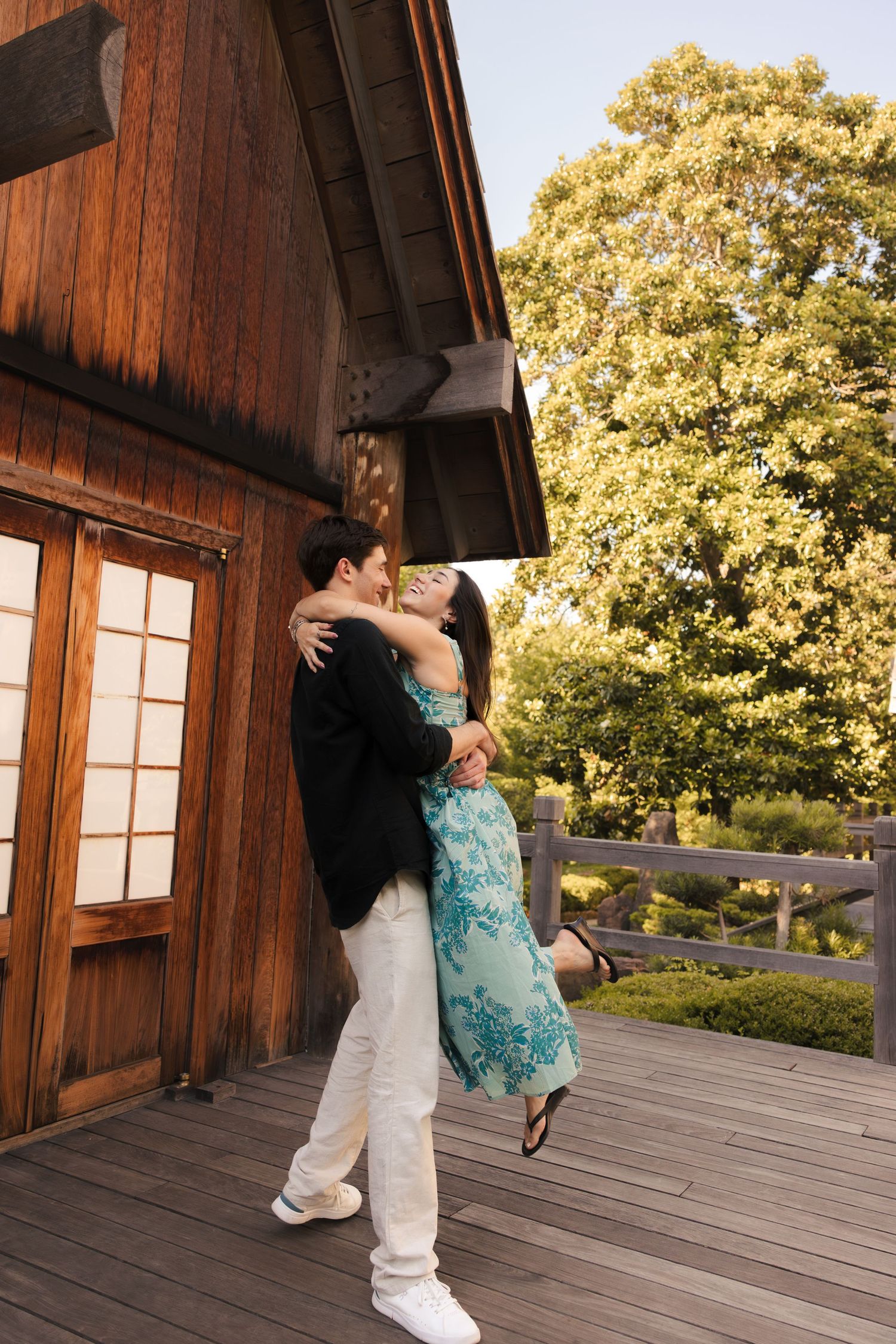 Couple embracing and kissing on a wooden deck outside a rustic building, surrounded by lush green trees.