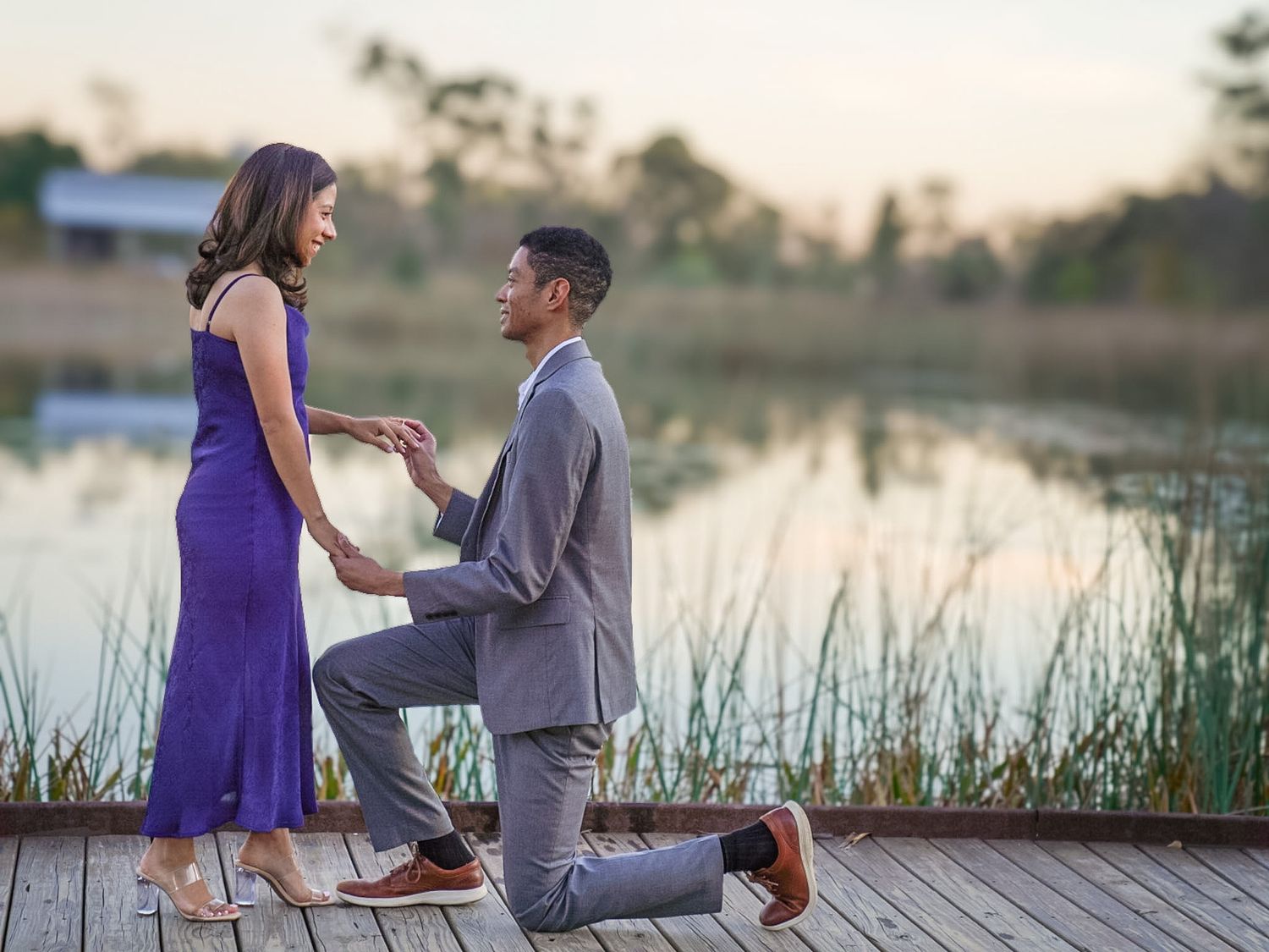 Man proposing to woman on wooden dock by lake at sunset, both dressed elegantly in purple and gray.