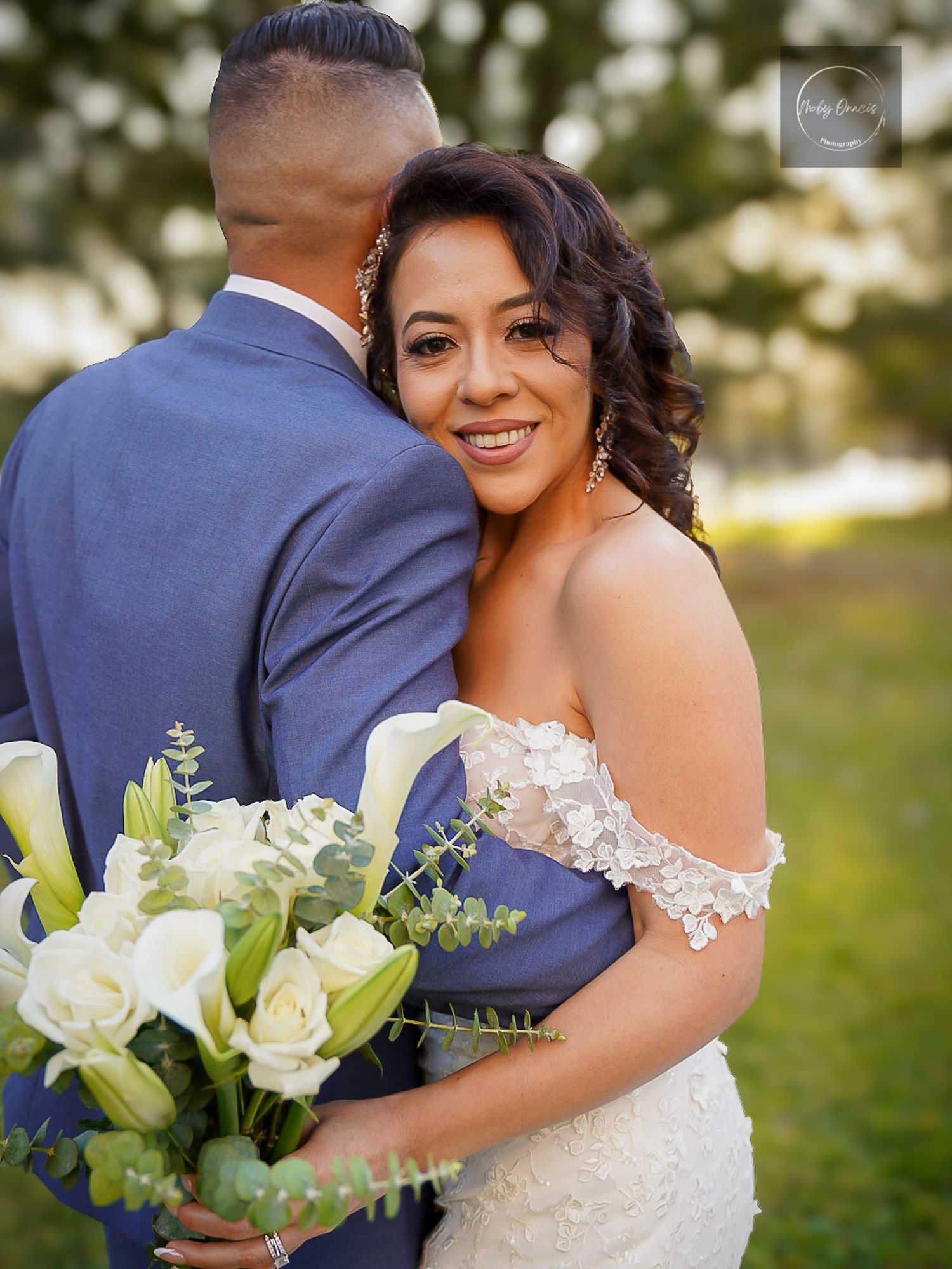Bride in lace off-shoulder gown holds white calla lily bouquet, embracing groom in blue suit outdoors.