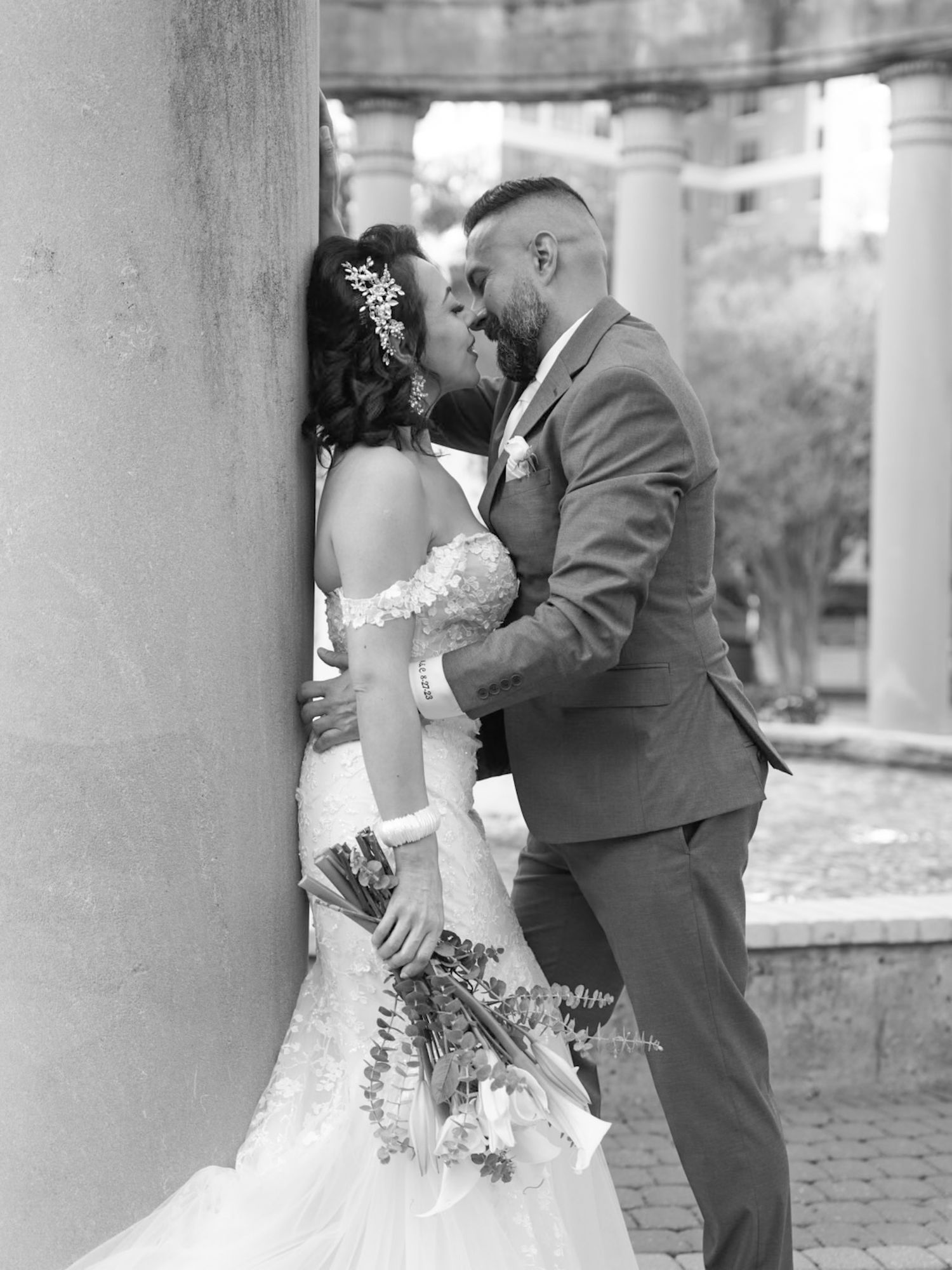 Black and white photo of a bride and groom sharing a kiss beside a stone column, bride holding a bouquet.