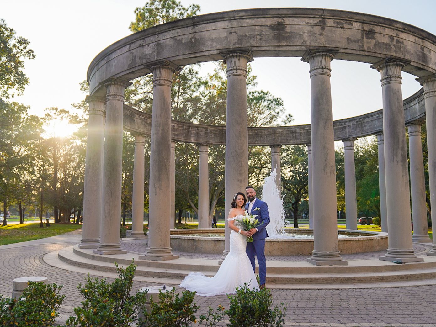 Bride and groom pose under a grand stone colonnade at sunset in a lush park setting.