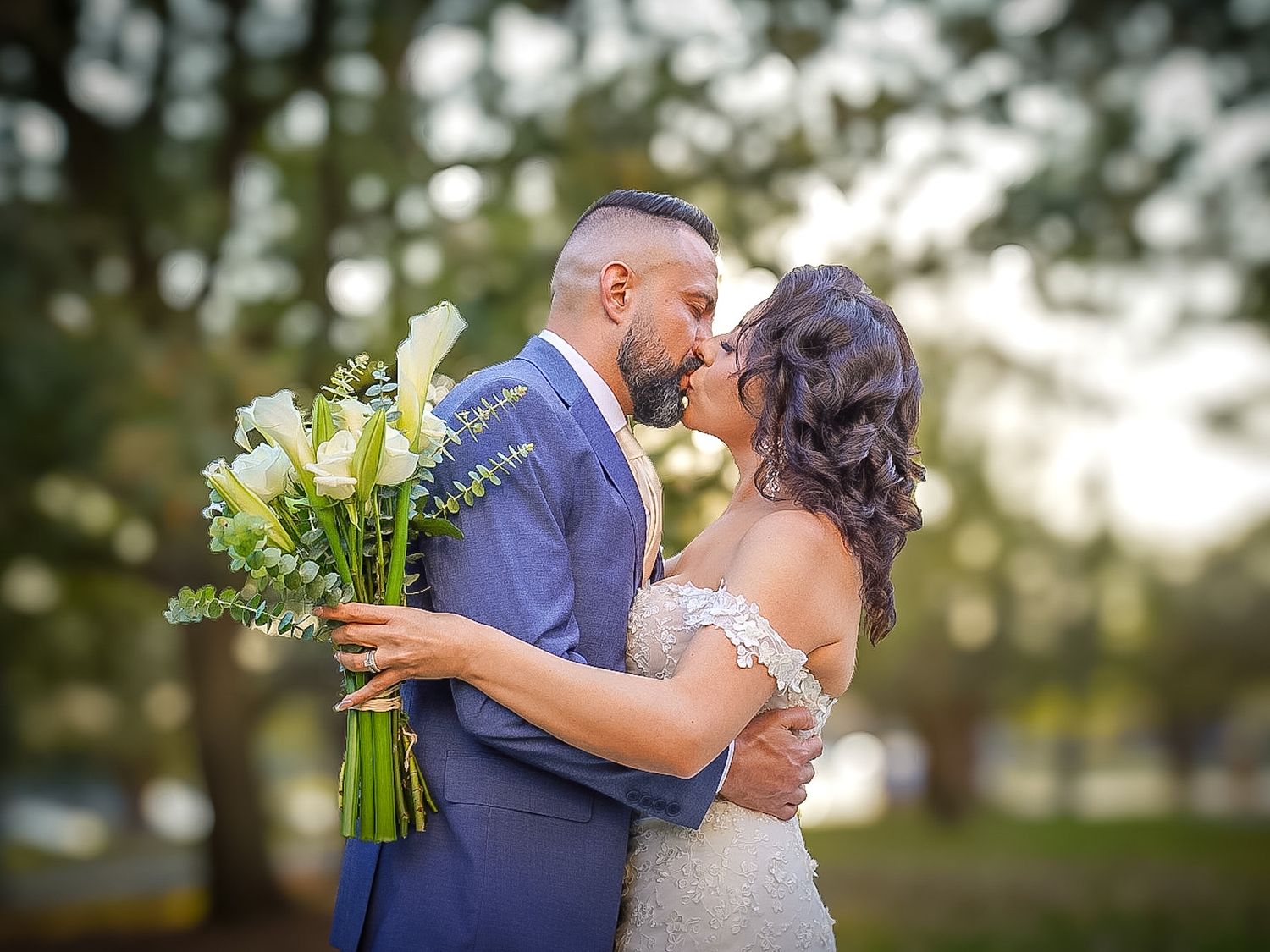 Bride and groom share a romantic kiss outdoors, bride holding white calla lily bouquet, groom in blue suit.