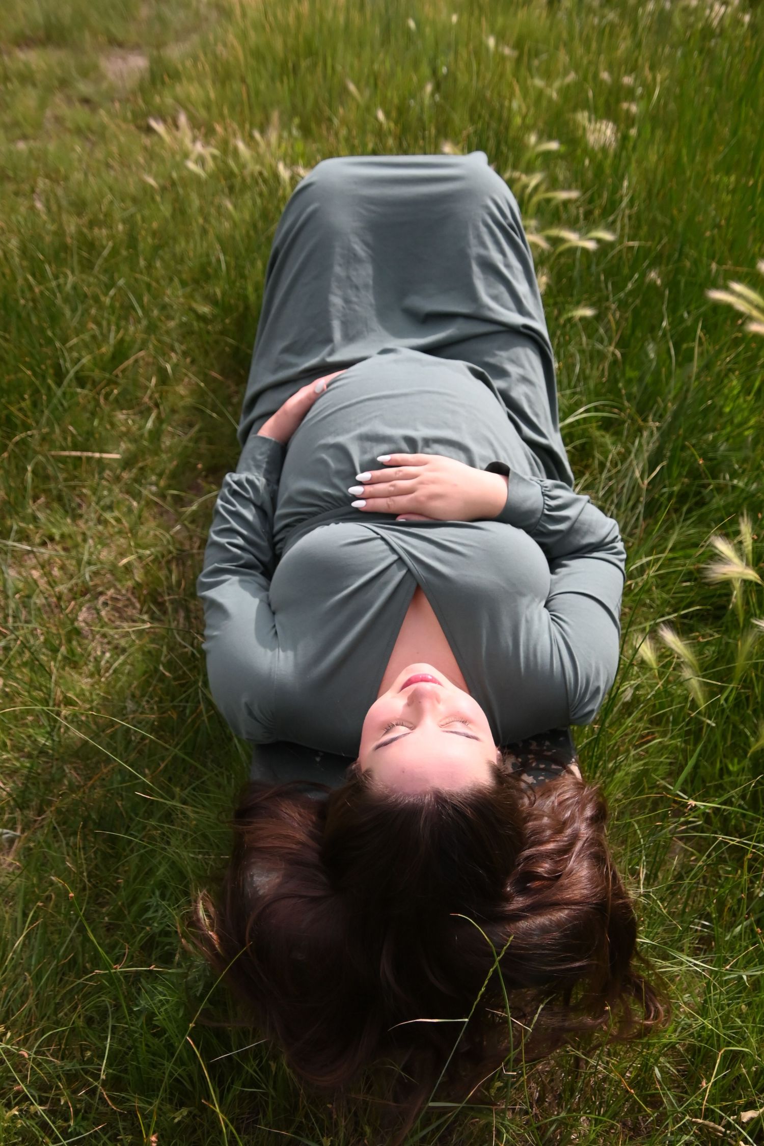 Pregnant woman in a gray dress lying in tall grass with their hands clasped over their stomach on a sunny day.