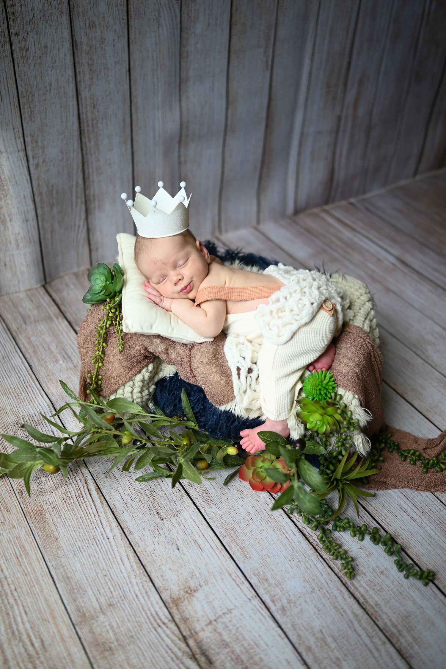 Peaceful newborn portrait with crown prop and greenery on rustic wooden floor.