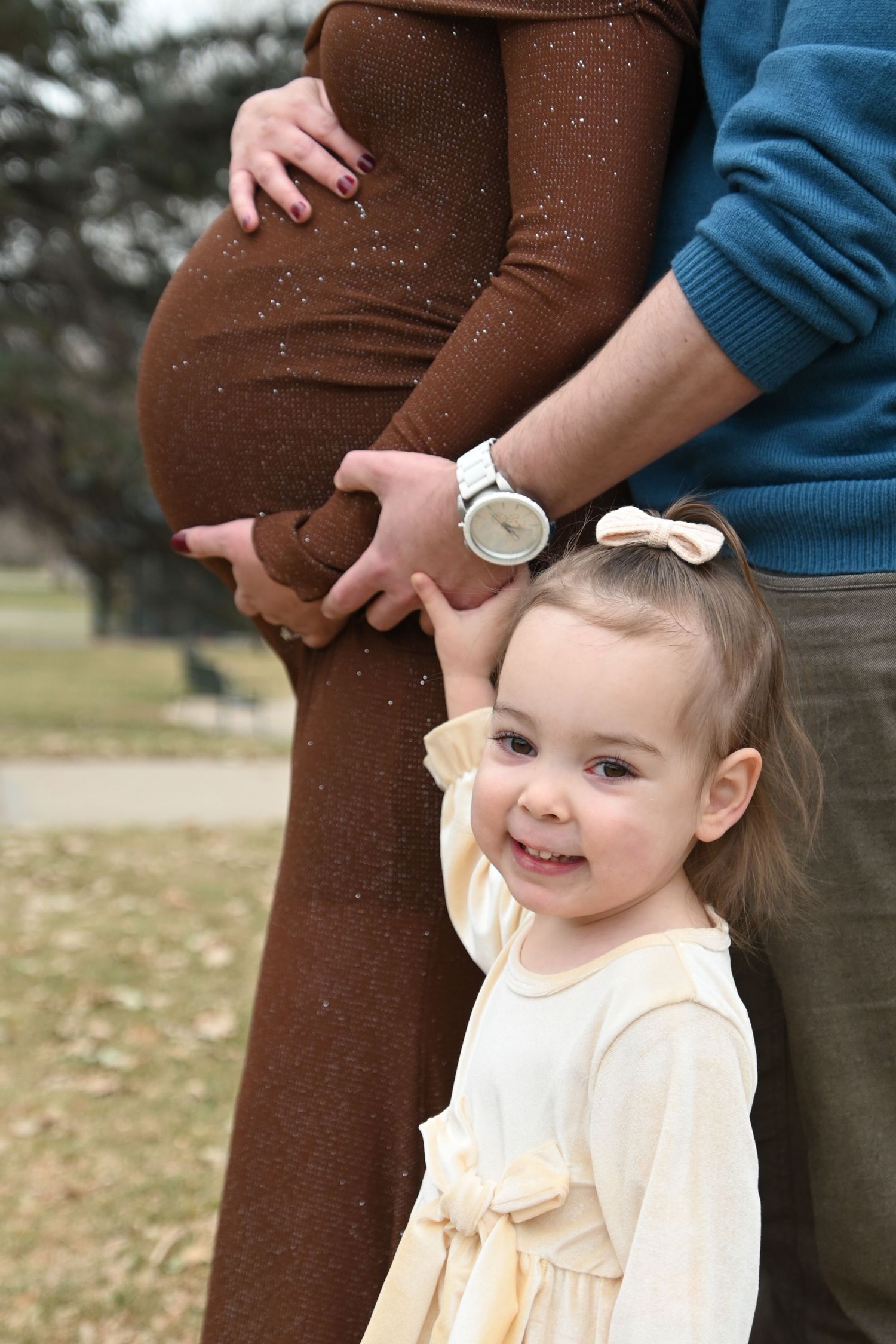 A little girl in a cream dress stands beside her pregnant mother wearing brown pants and someone in a blue sweater.