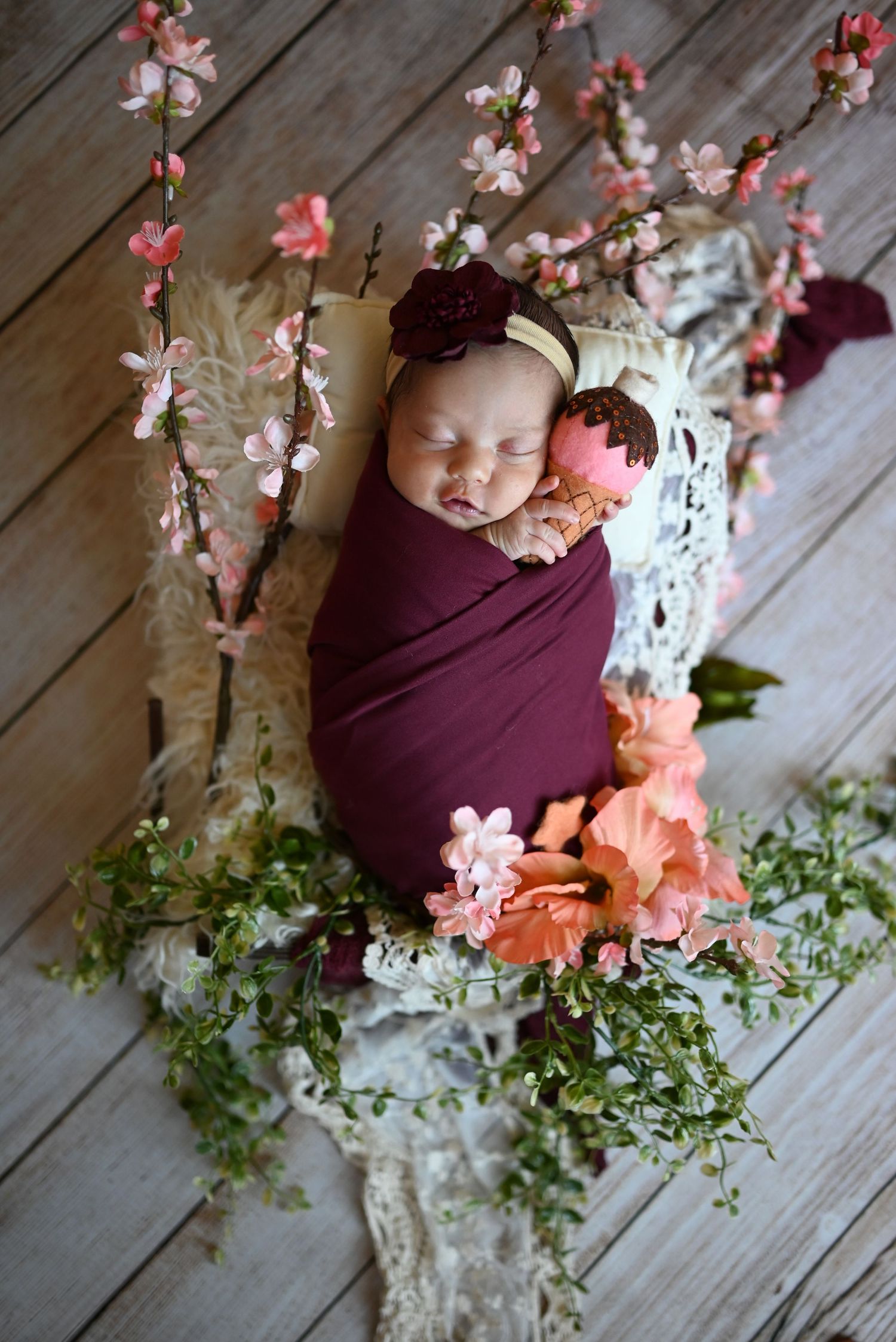 A cozy newborn photography setup with burgundy wrap, delicate pink cherry blossoms, and greenery on wooden floorboards.