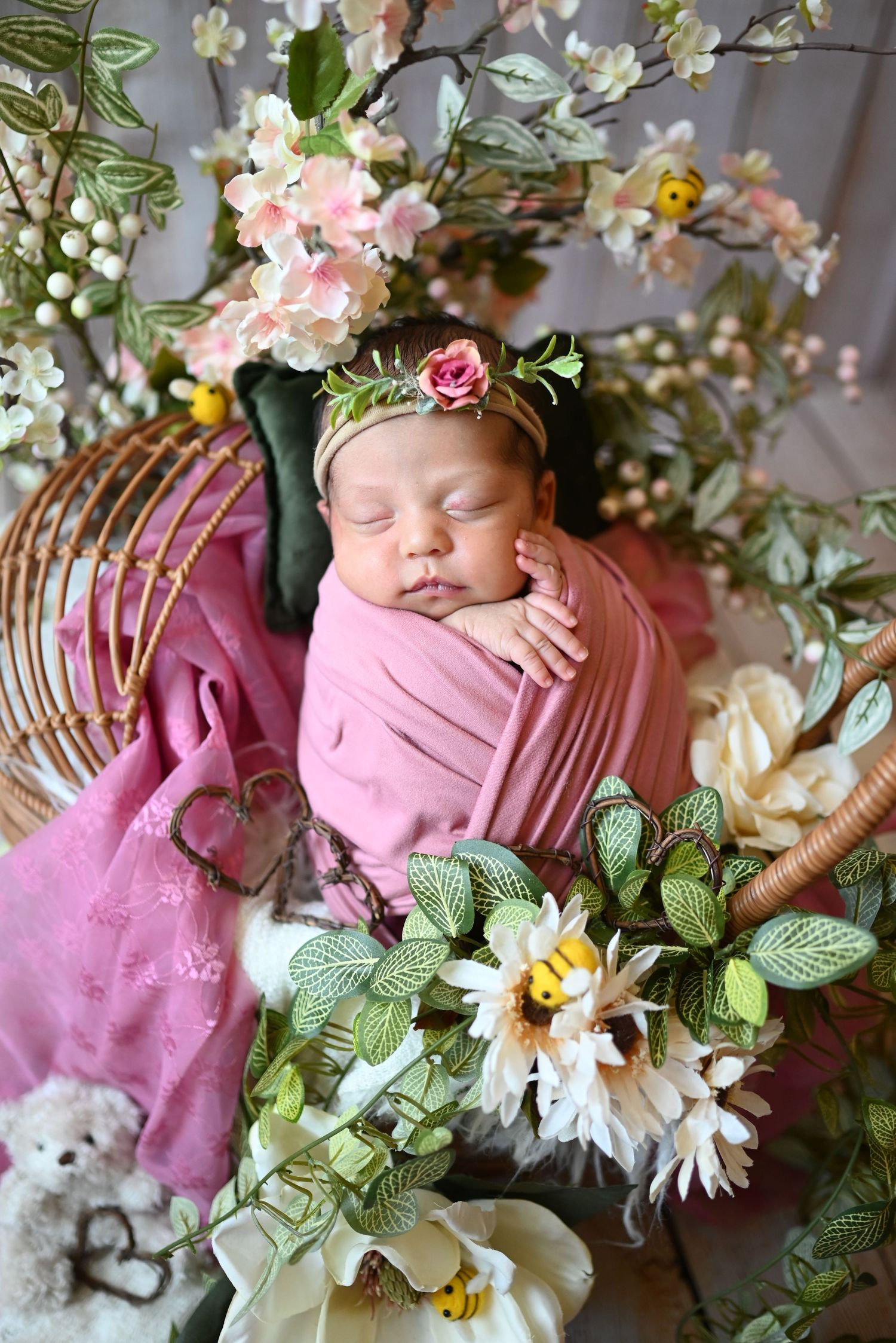 Newborn photography setup with pink wrap and floral arrangements on wicker chair.