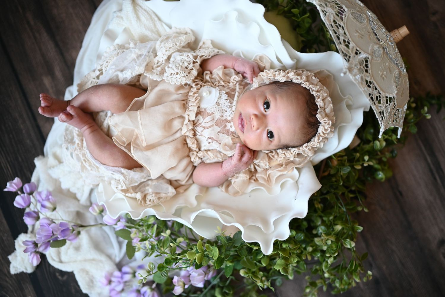 Baby in vintage lace dress posed with purple flowers on dark wooden background.