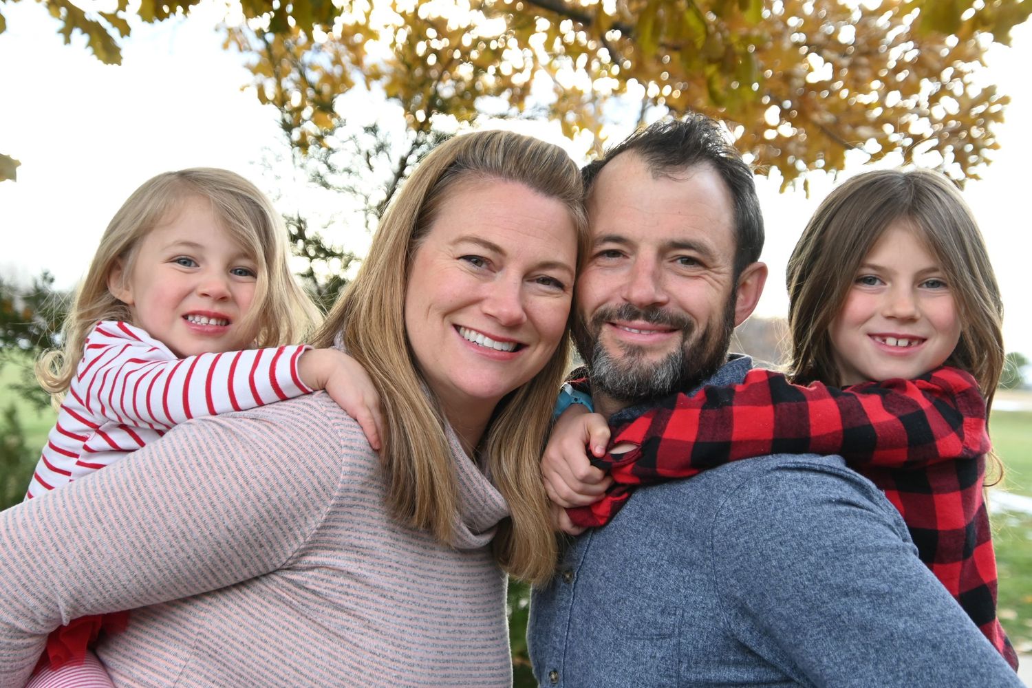 Family of four poses together outdoors with autumn foliage in background during fall photo session.