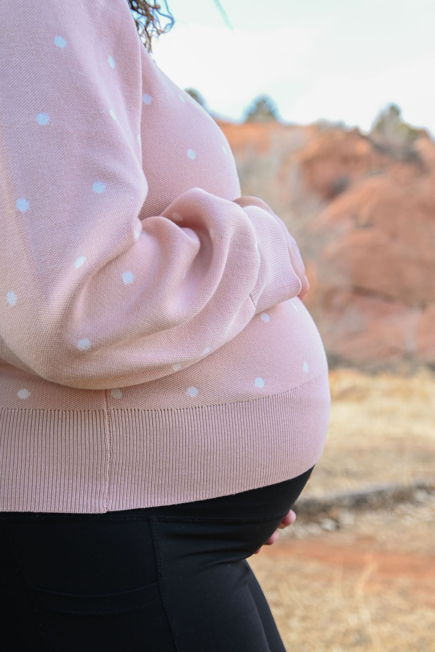 A pregnant person wearing a pink polka dot sweater and black pants stands against a desert landscape background.
