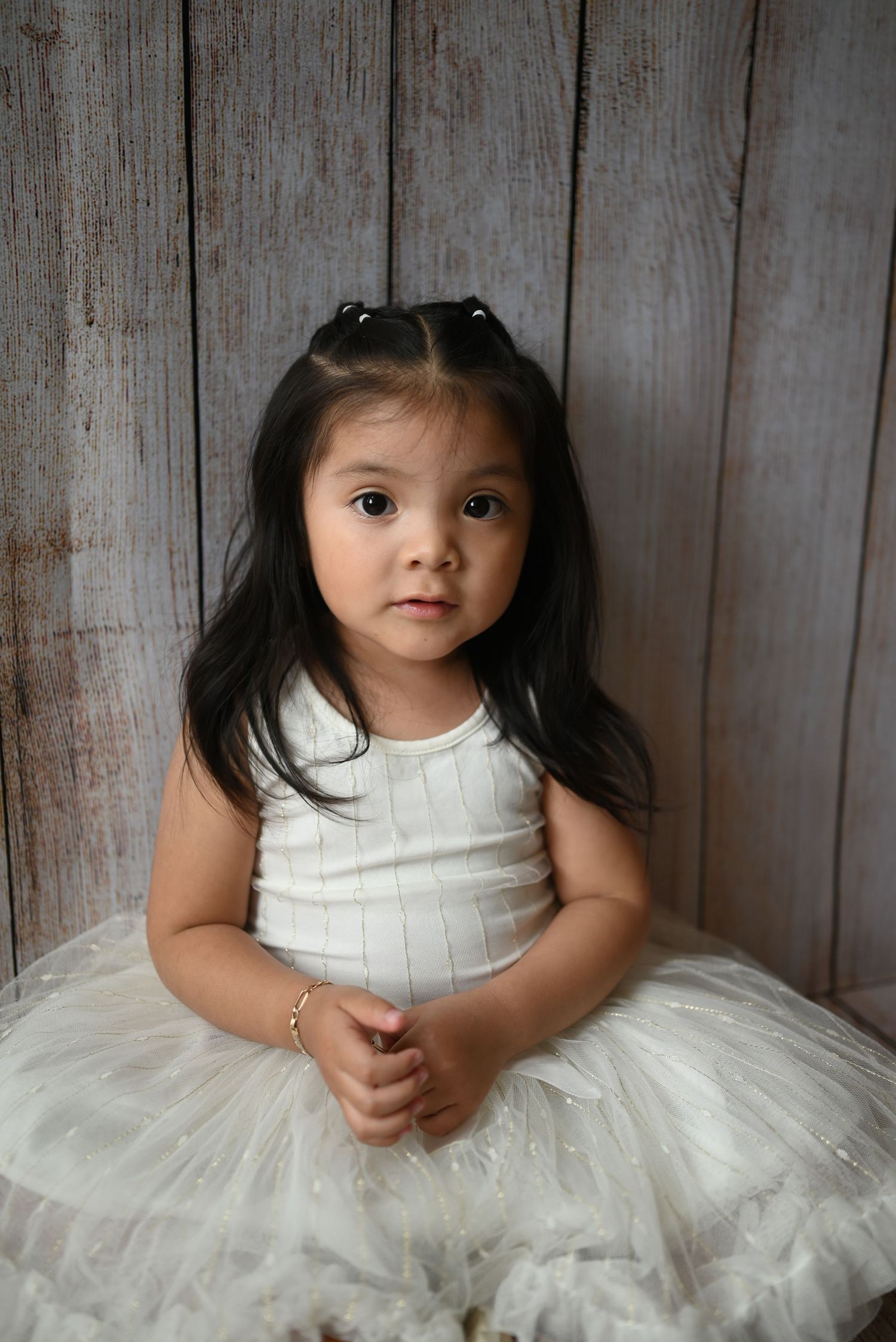 Young child wearing a white tutu and tank top poses against rustic wooden background.