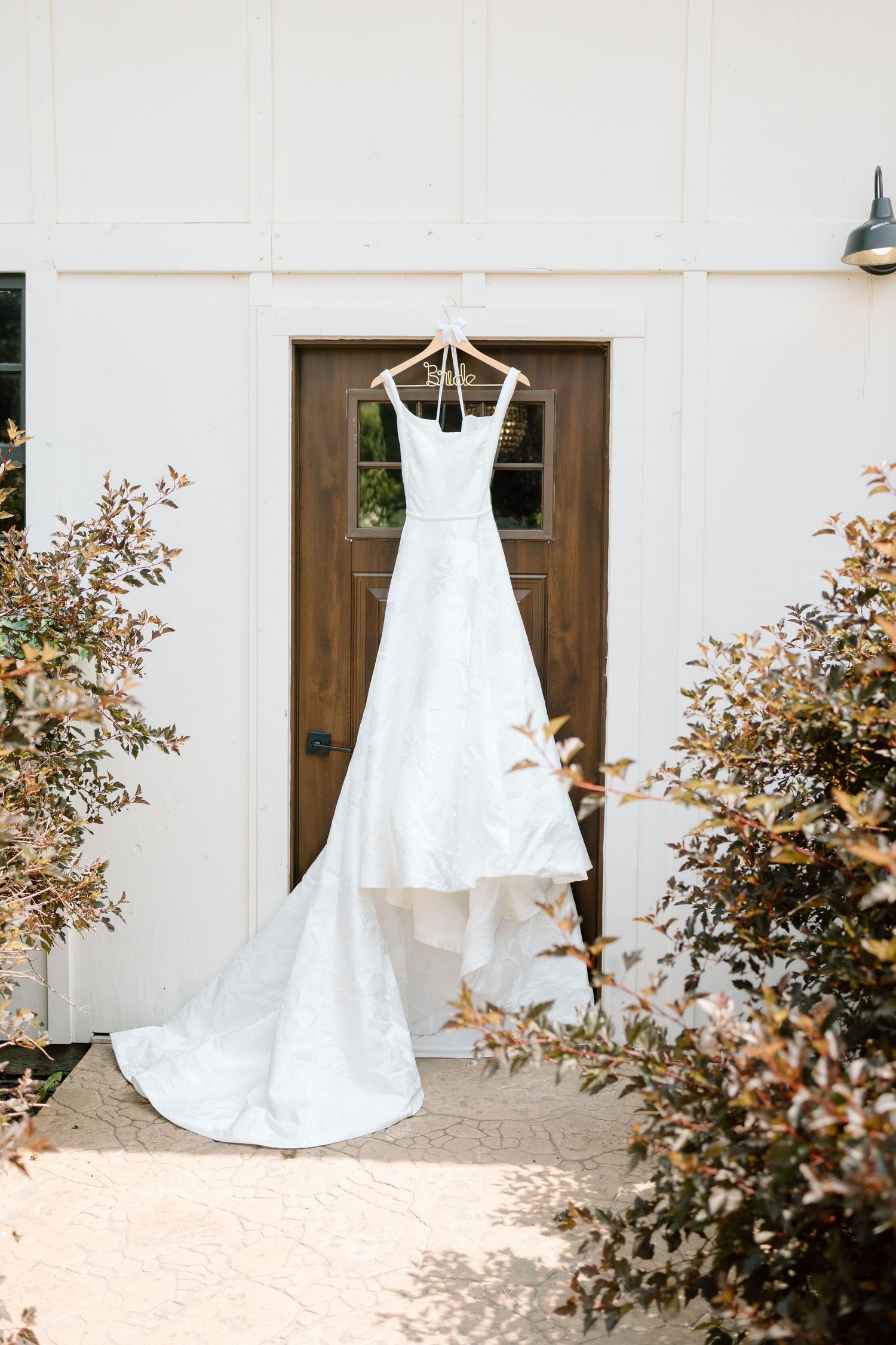A white wedding dress hangs elegantly against a beige wall with decorative autumn foliage in the foreground.