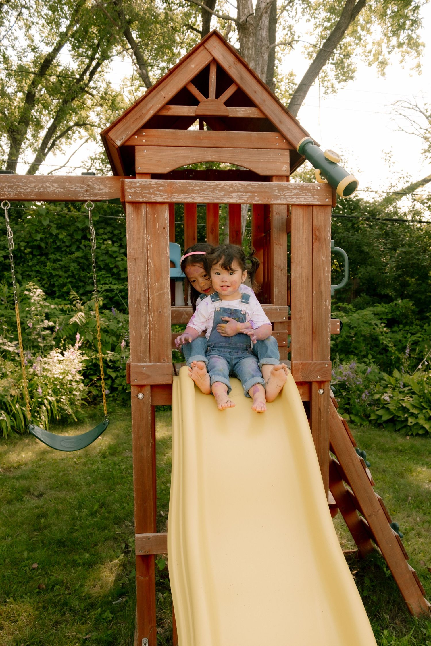 Two children playing on a wooden backyard playset, sliding down a yellow slide surrounded by lush green trees.