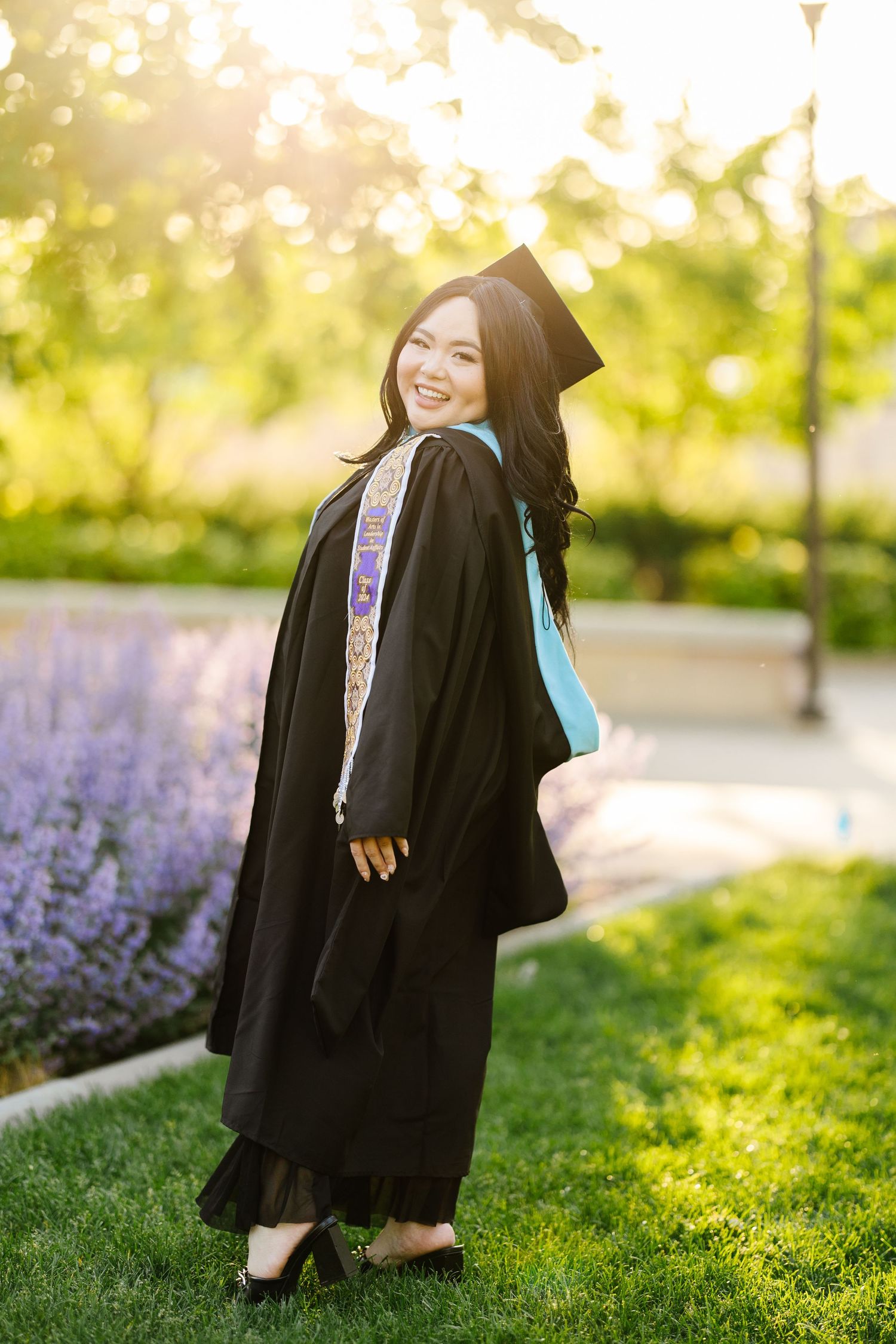 Graduate in traditional academic regalia poses joyfully in garden with blooming purple flowers at sunset.