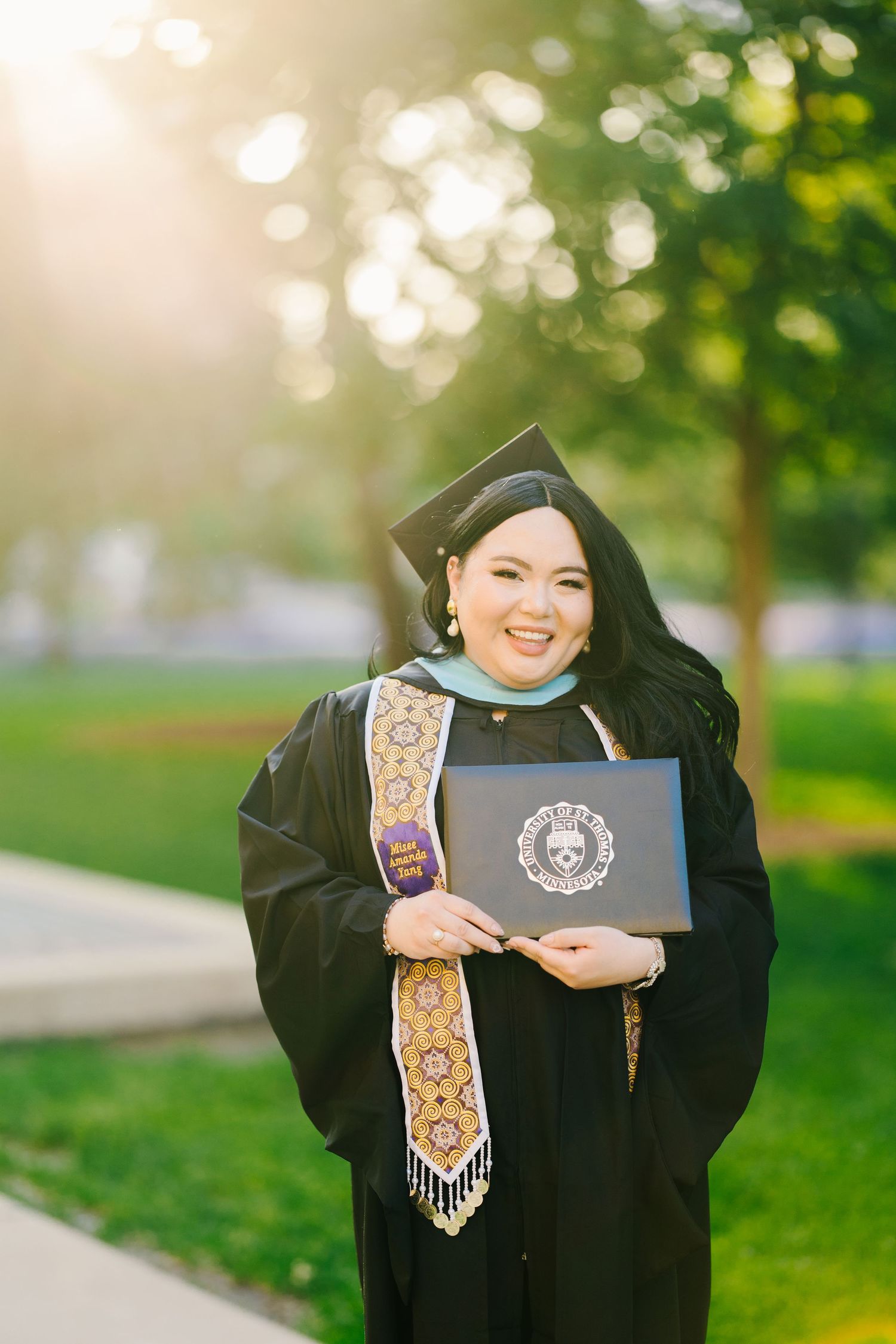 A graduate in traditional academic regalia holds a diploma on a sunny day with bokeh-lit trees in the background.