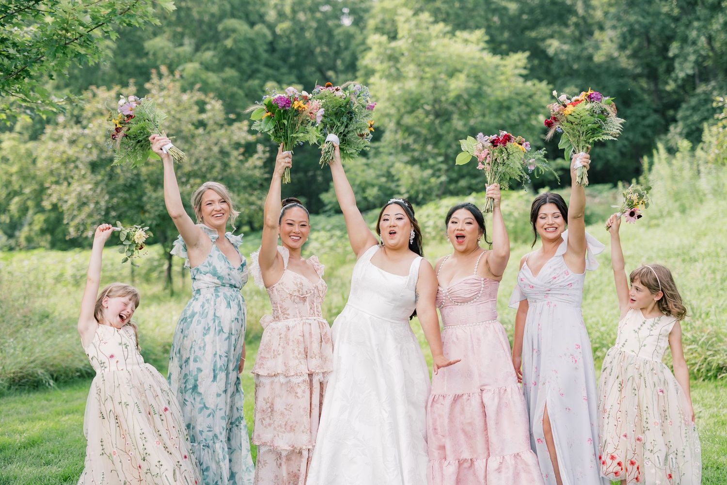 Wedding party in pastel dresses celebrating joyfully with bouquets raised in an outdoor setting.