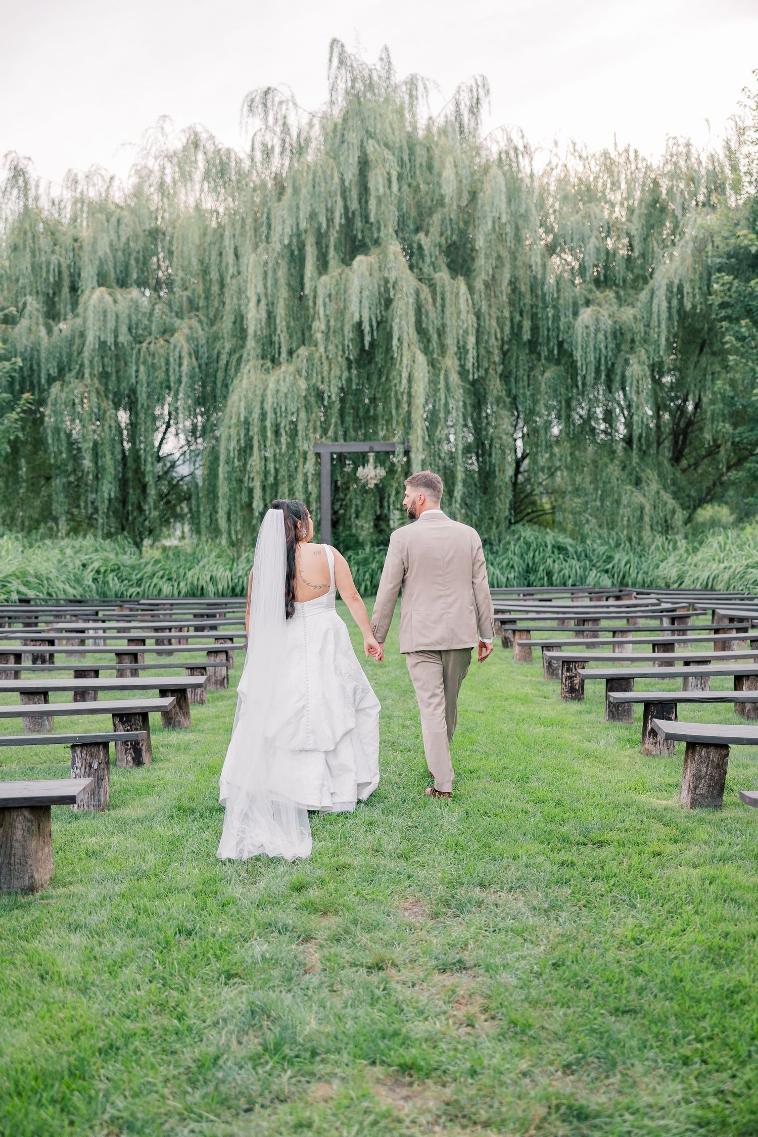 Wedding couple walking through an outdoor ceremony space with wooden benches under weeping willow trees.