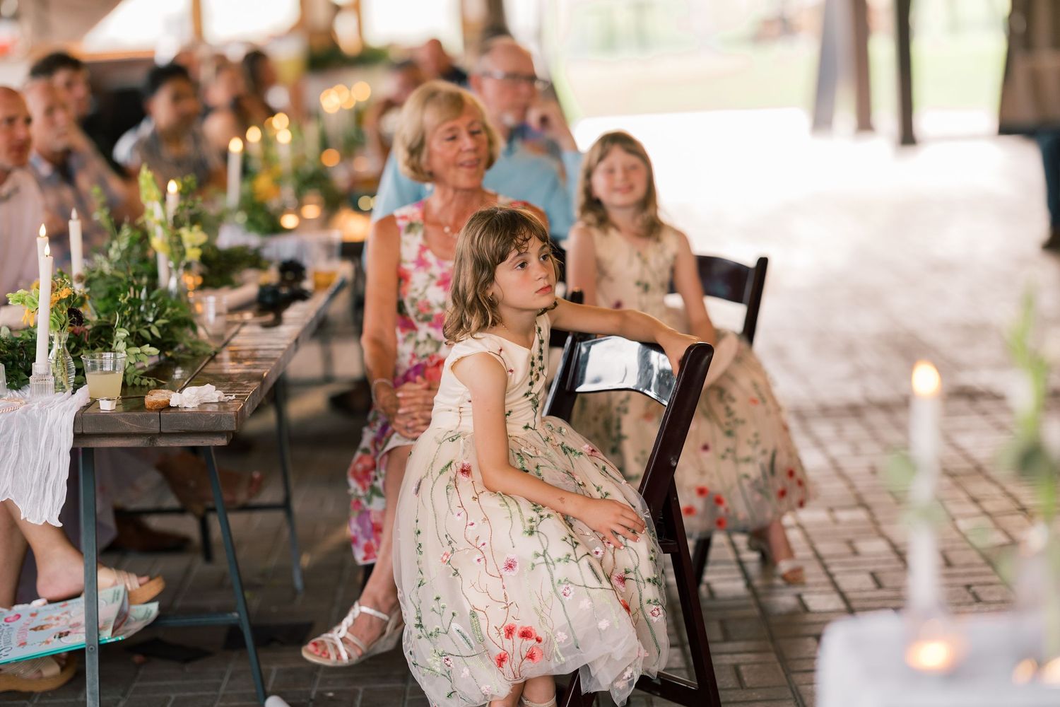Wedding guests seated at a reception table outdoors with decorative candles and greenery centerpieces.