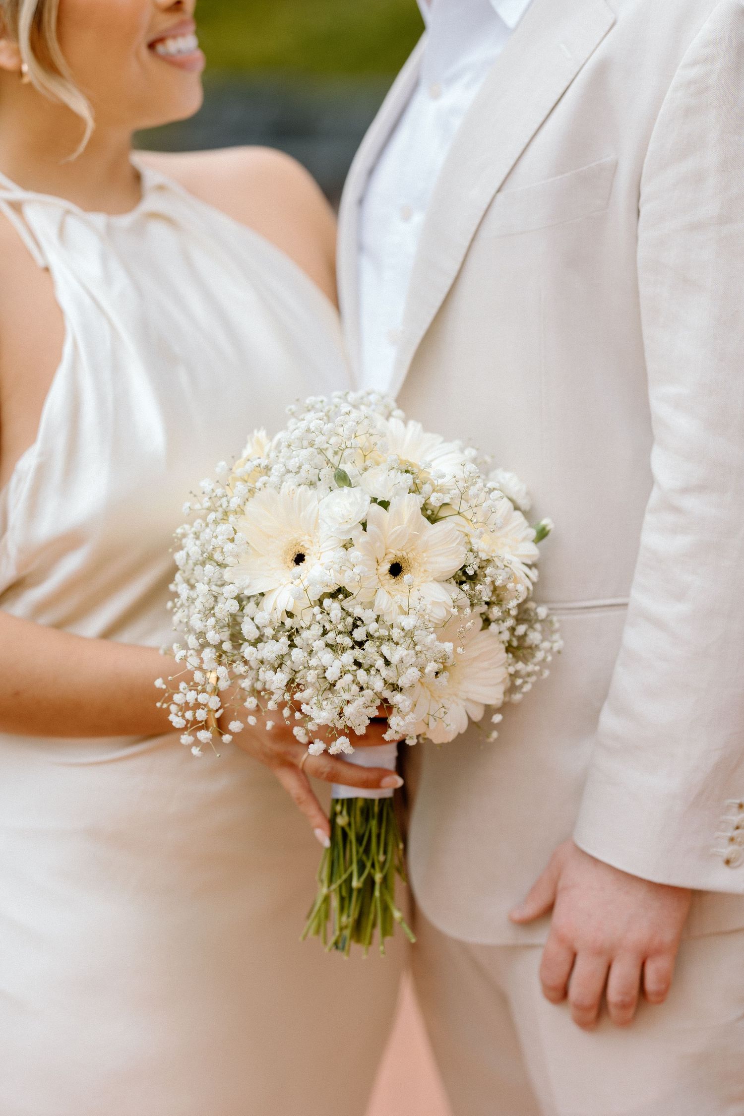 A delicate white wedding bouquet with daisies and baby's breath held against a white dress and suit.