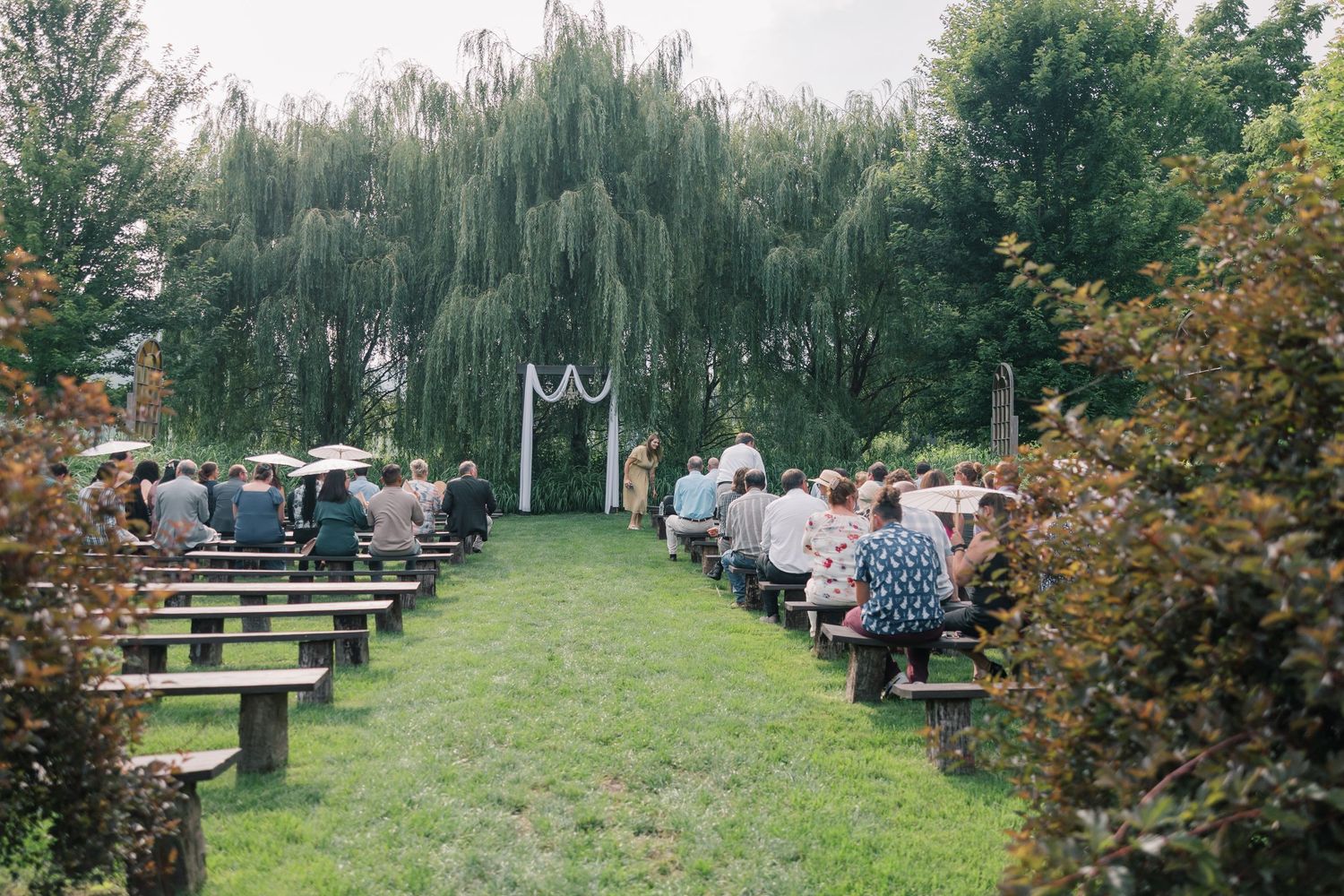 Outdoor wedding venue with willow trees and picnic tables set up on green grass.