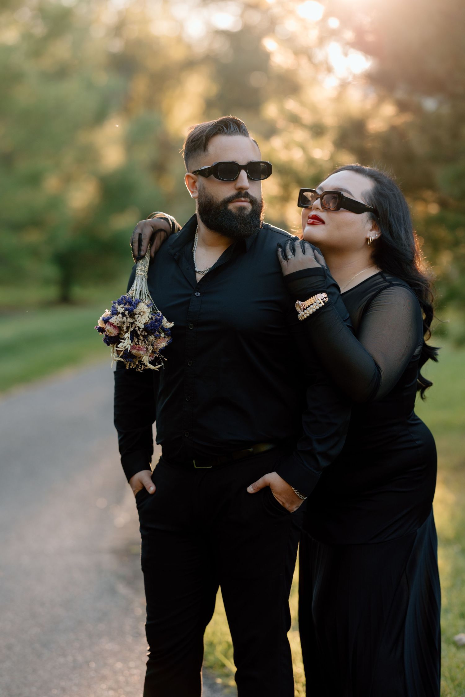 A stylish couple in matching black outfits poses together on a path with autumn trees in the background.