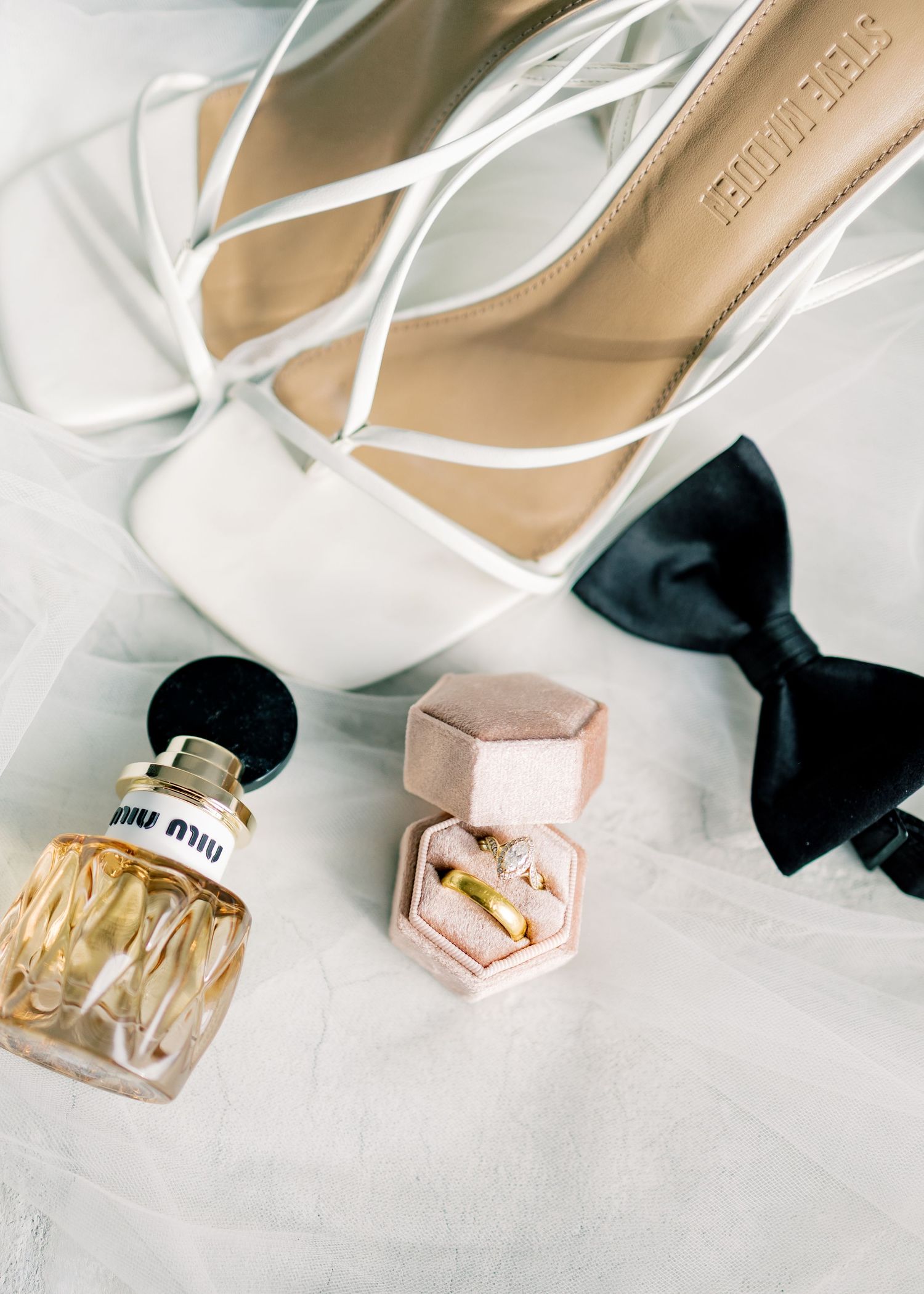 Wedding accessories including white strappy heels, perfume bottle, black bow tie and pink ring box arranged on white fabric.