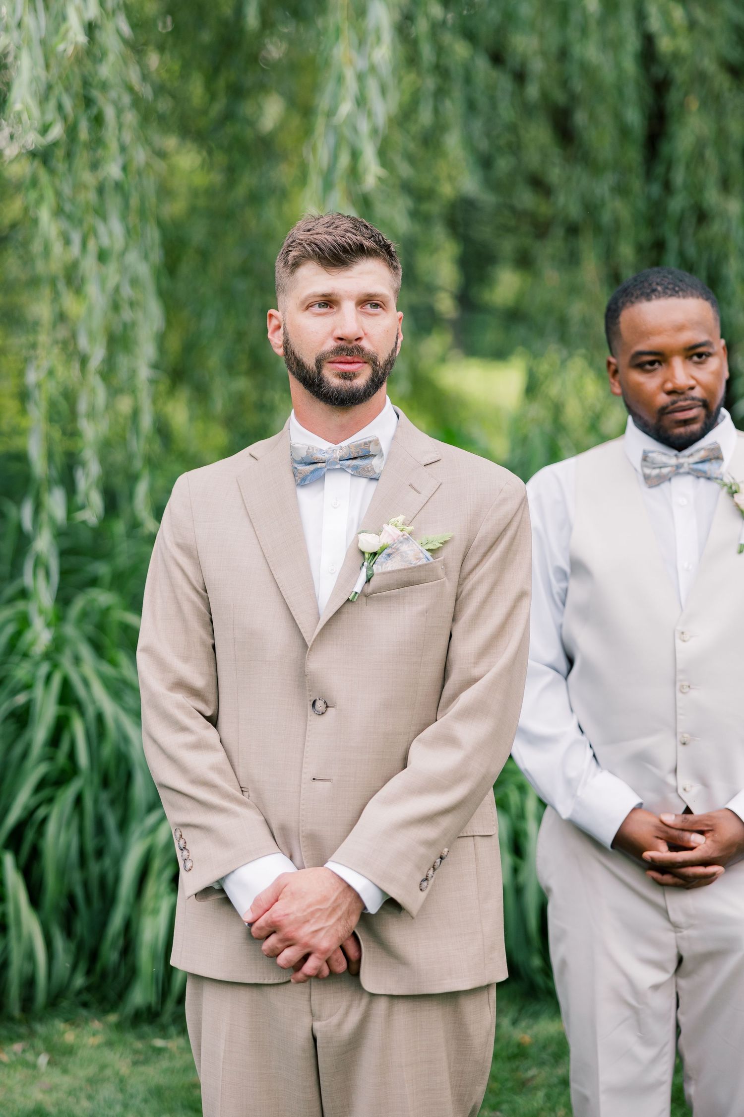 Wedding party members in tan and light gray suits standing together outdoors near willow trees.
