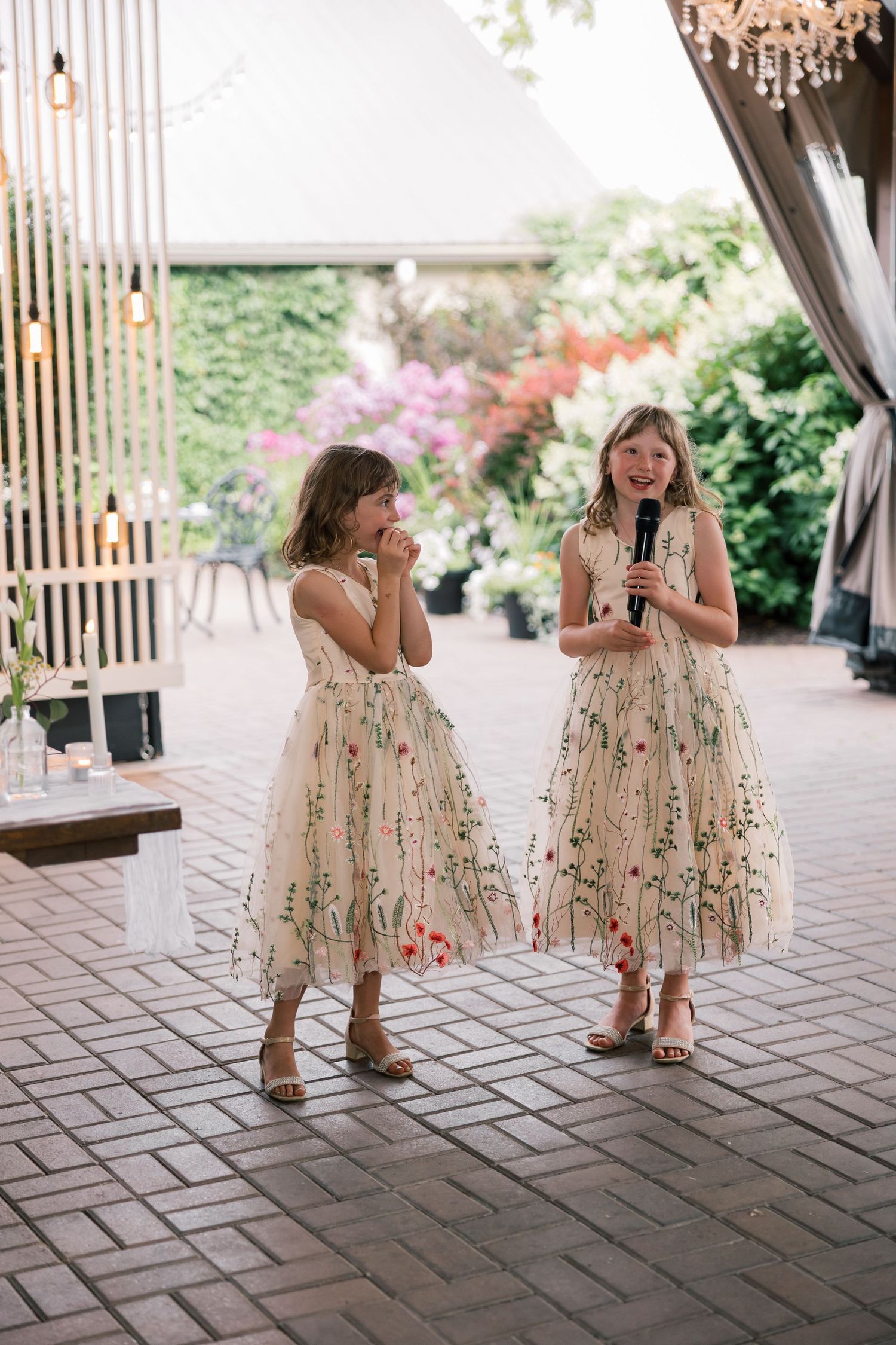 Two young attendants in floral dresses dancing on a brick patio at a wedding reception.