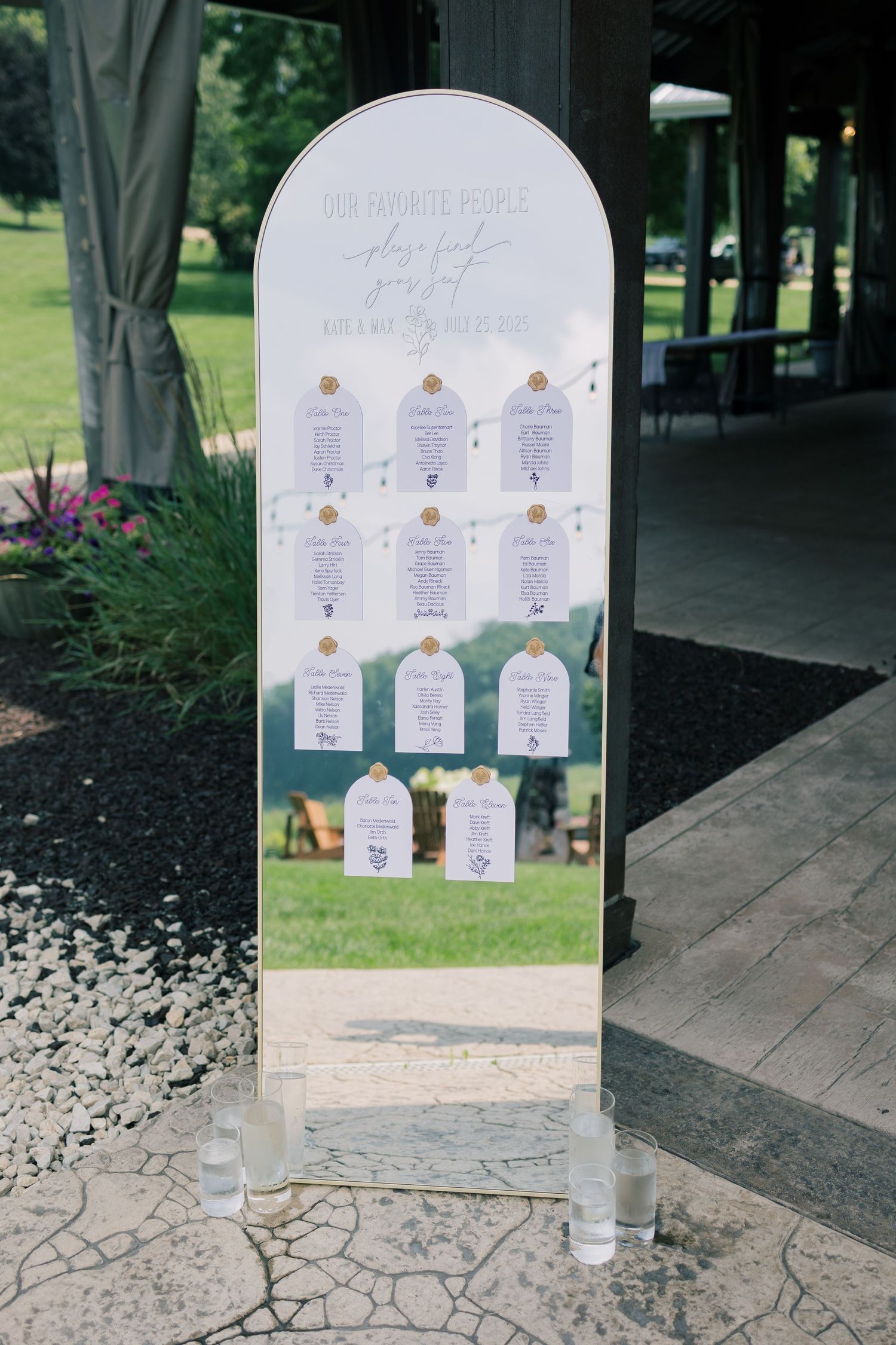 A white seating chart display board with elegant wedding table assignments arranged in a decorative pattern.