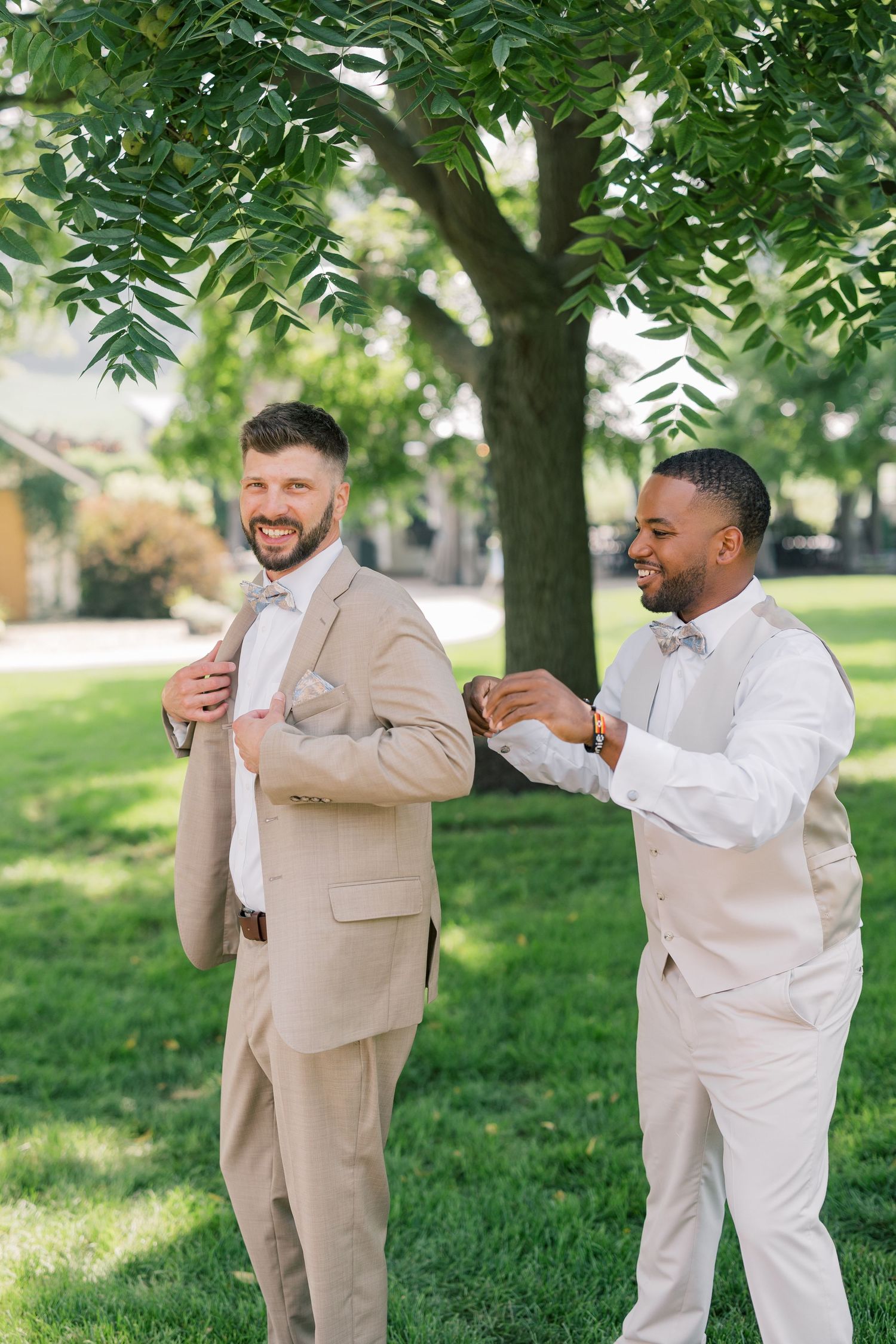 Two men in contrasting beige and white suits share a playful moment outdoors under green trees.