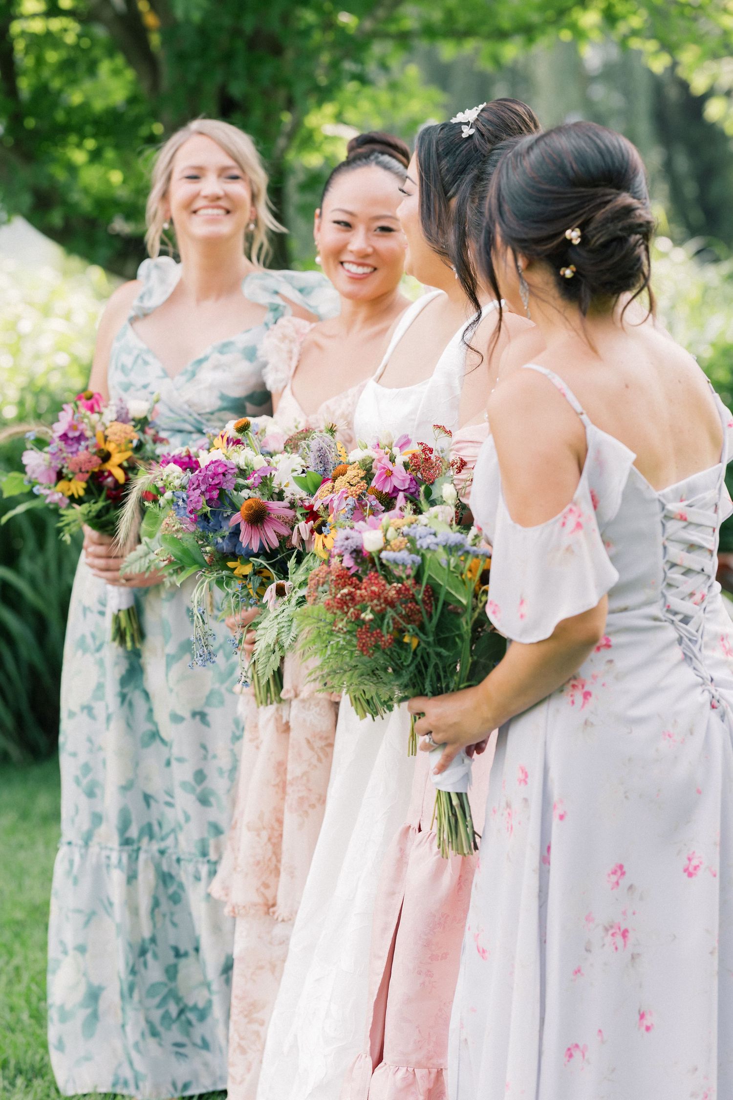 Bridesmaids in pastel floral dresses hold vibrant wildflower bouquets outdoors.