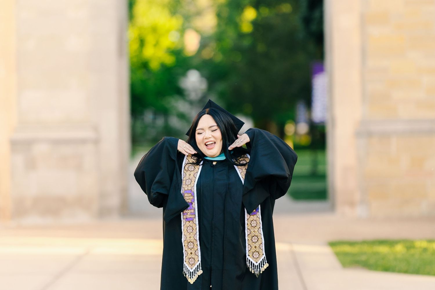 Graduate in black cap and gown poses joyfully with decorated stole during outdoor celebration.