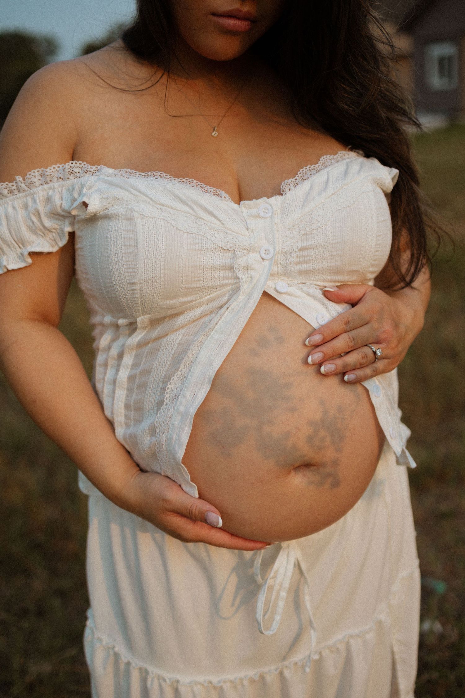 Pregnant woman in white off-shoulder dress holding baby bump outdoors, showing floral tattoo on stomach.