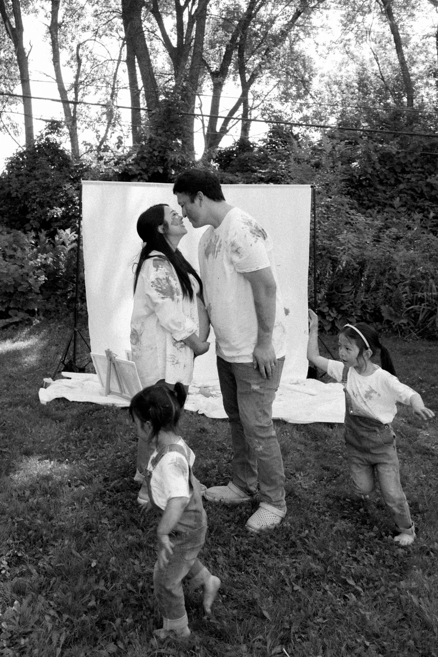 Black and white photo of couple kissing during outdoor family paint photo shoot with two young children playing nearby.