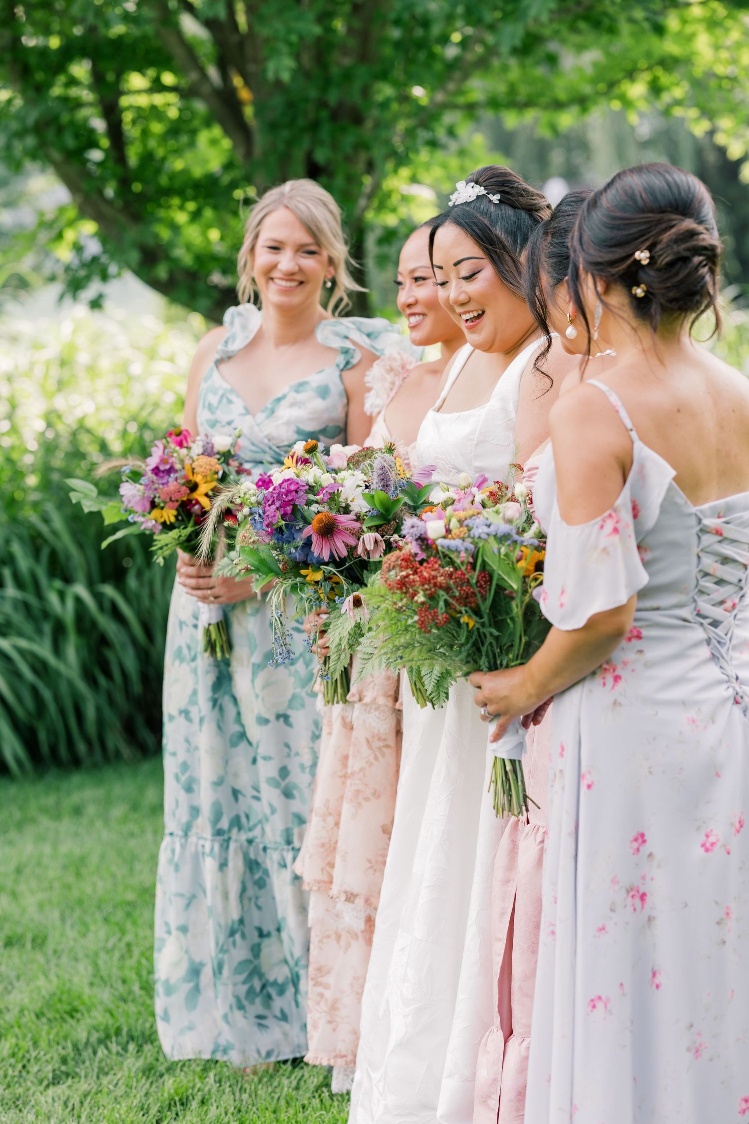 Bridesmaids in pastel floral dresses stand together holding colorful wildflower bouquets outdoors.