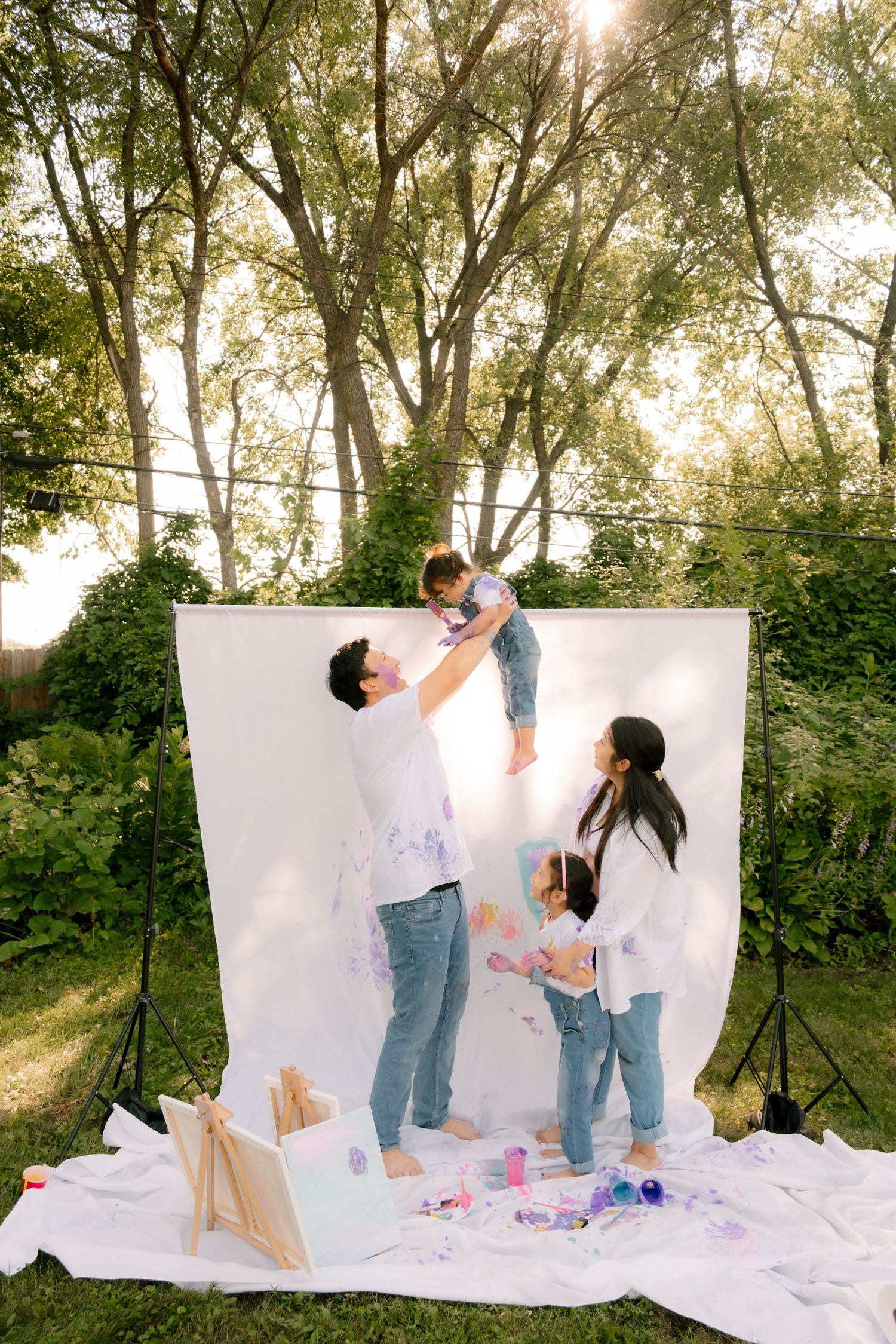 Family with paint-covered clothes posing outdoors against white backdrop, father lifting child while mother and daughter stand nearby.