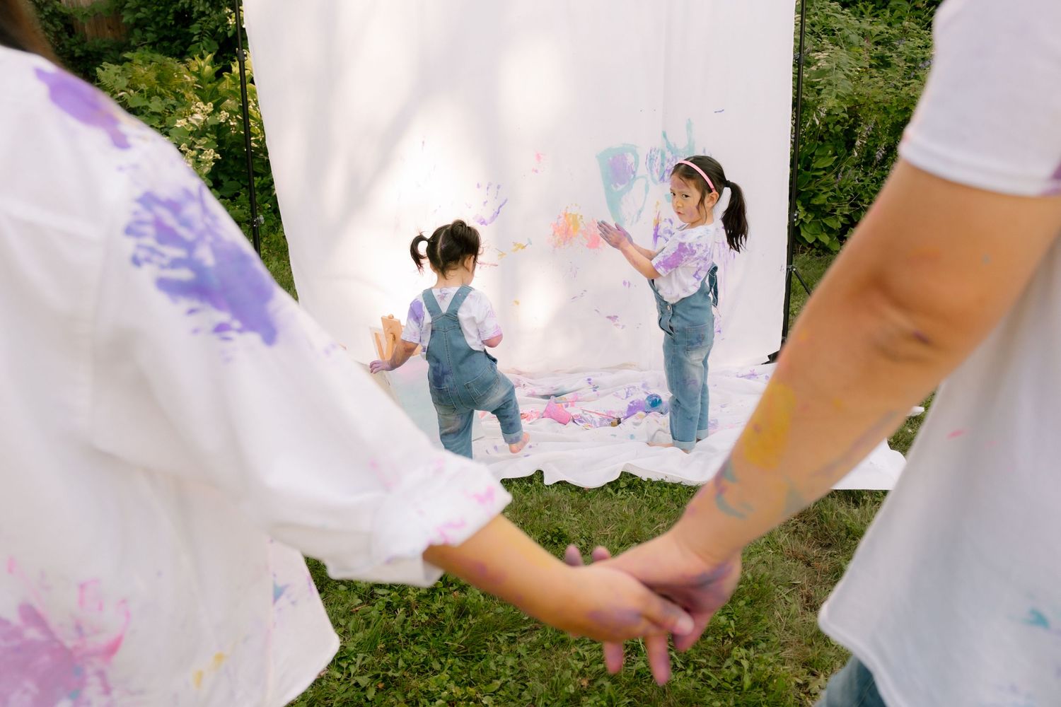 Two children painting on white canvas sheets outdoors while adults hold hands in the foreground.