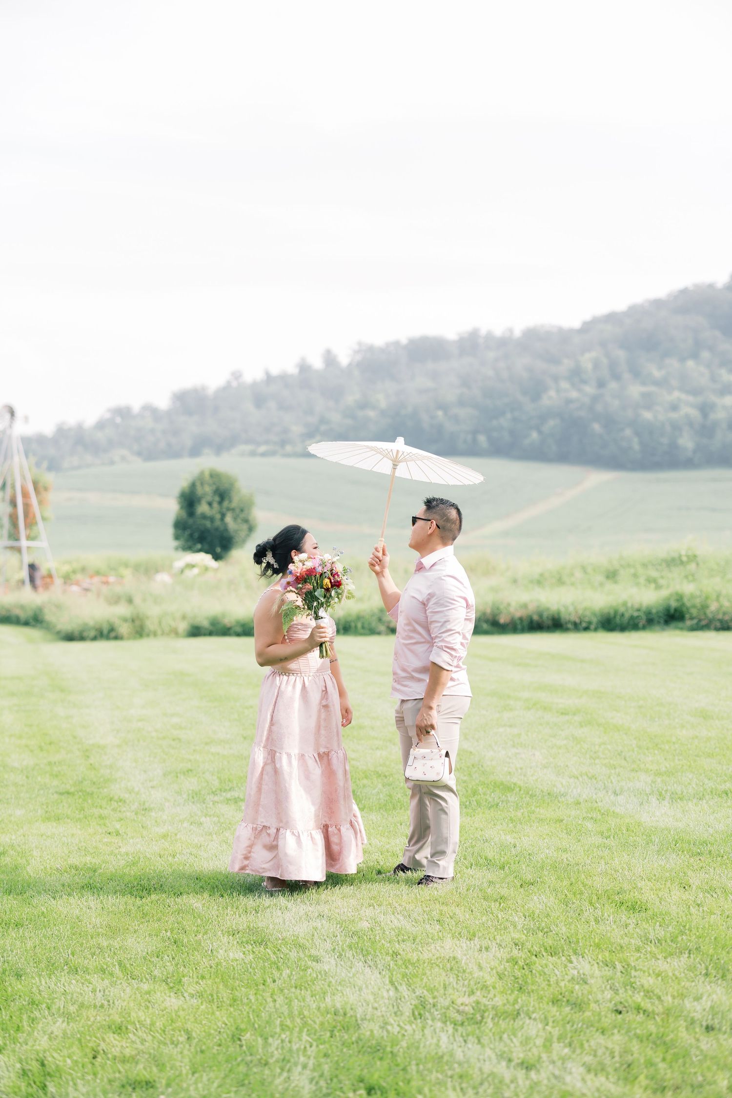 A couple in soft pink and beige outfits share a romantic moment in a lush green hillside meadow.
