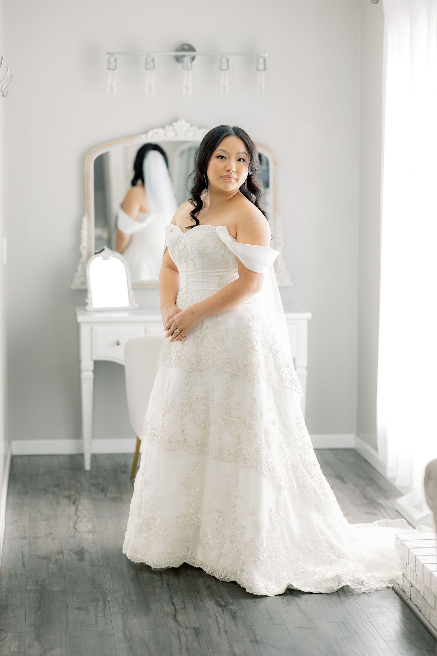 A wedding dress displayed in a bright dressing room with white vanity and mirror.