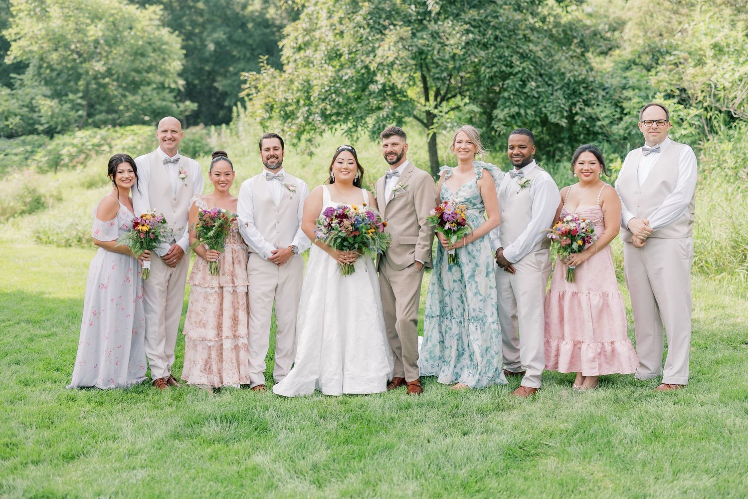 Wedding party poses together on grassy hillside wearing pastel colored dresses and suits.