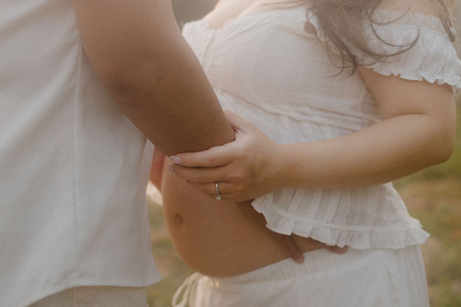 Pregnant woman in white dress holding her baby bump with partner outdoors in soft natural light.