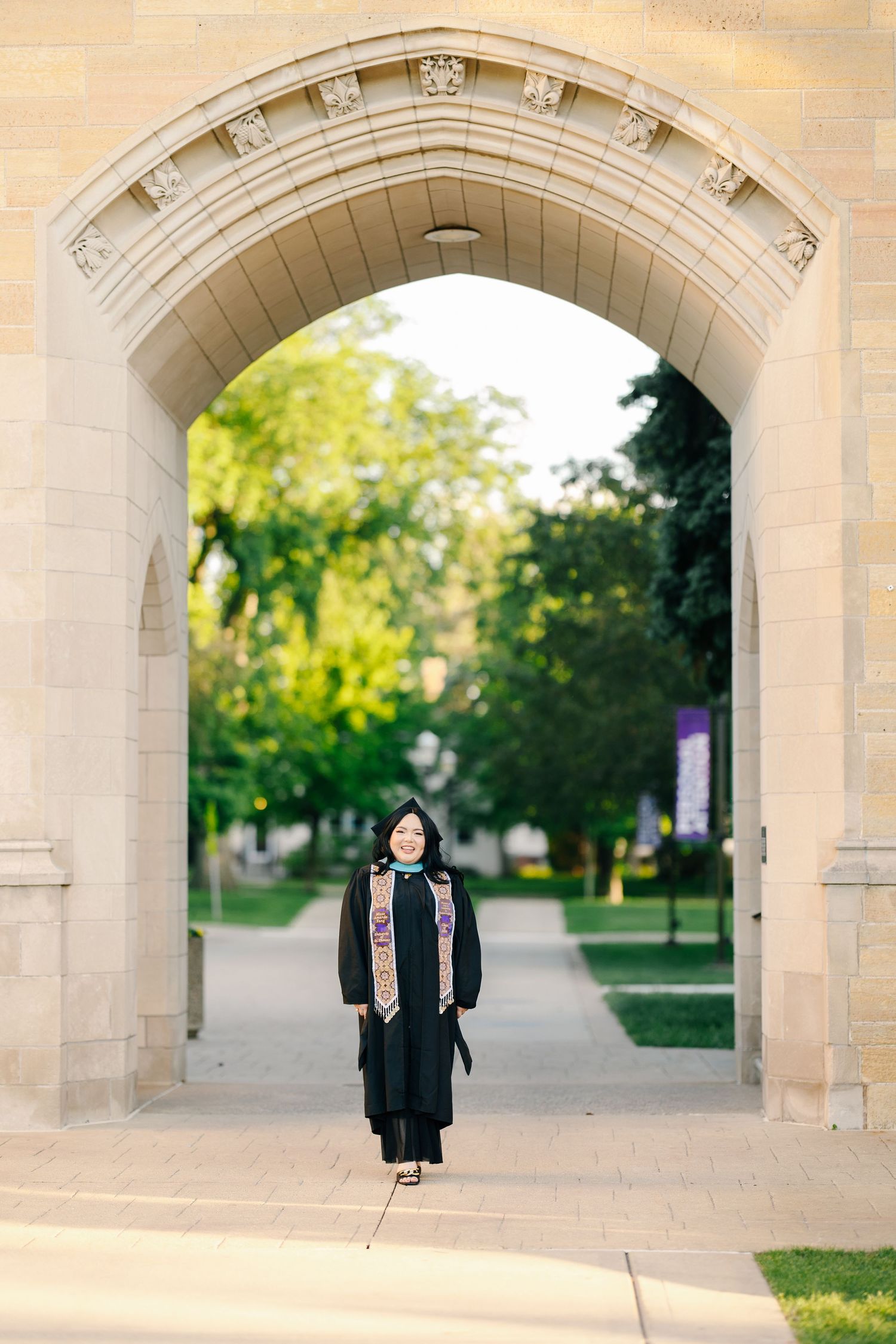 Graduate stands under a grand stone archway on a university campus walkway.