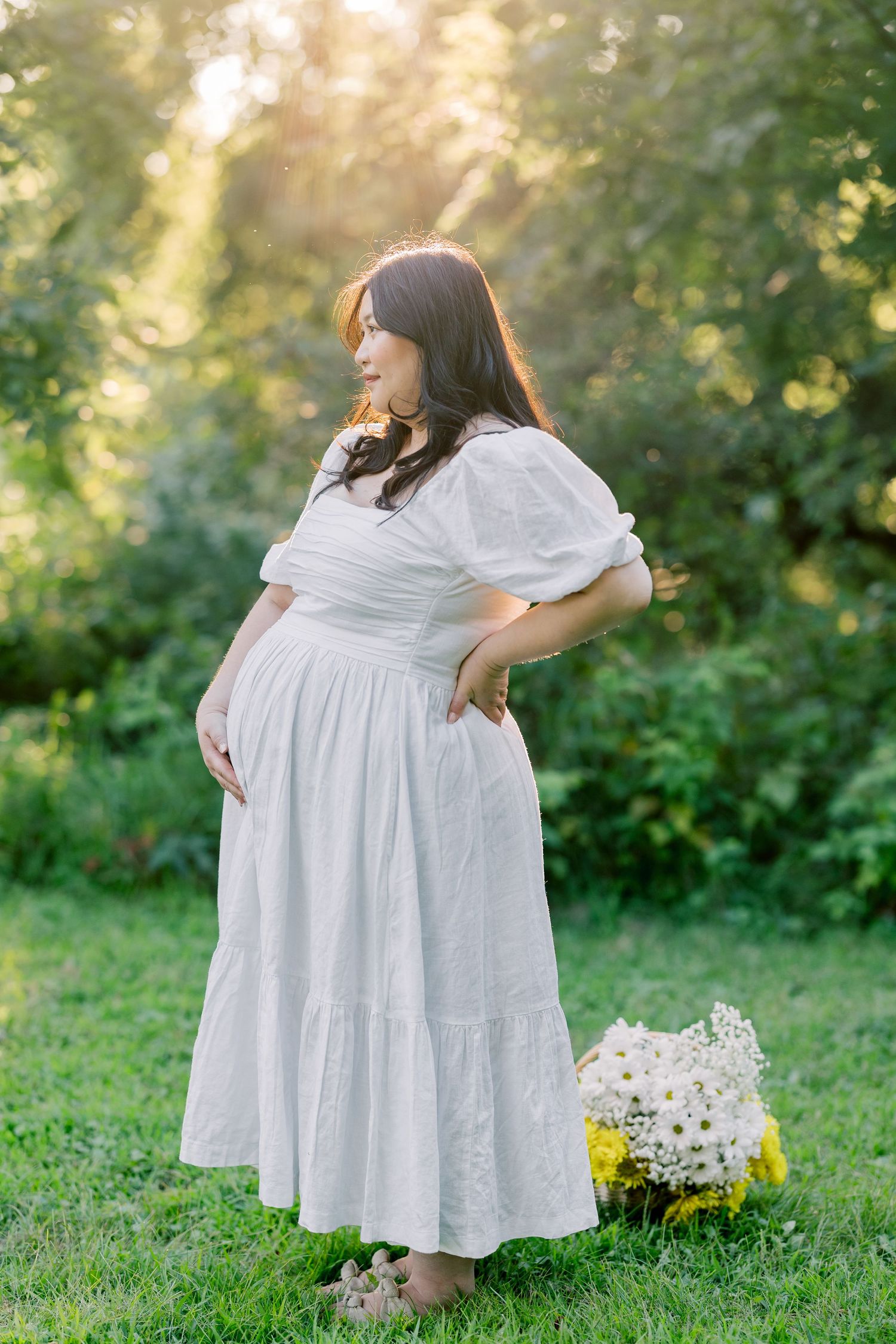A pregnant figure wearing a flowing white cottagecore dress poses in a sunlit garden with flowers at their feet.