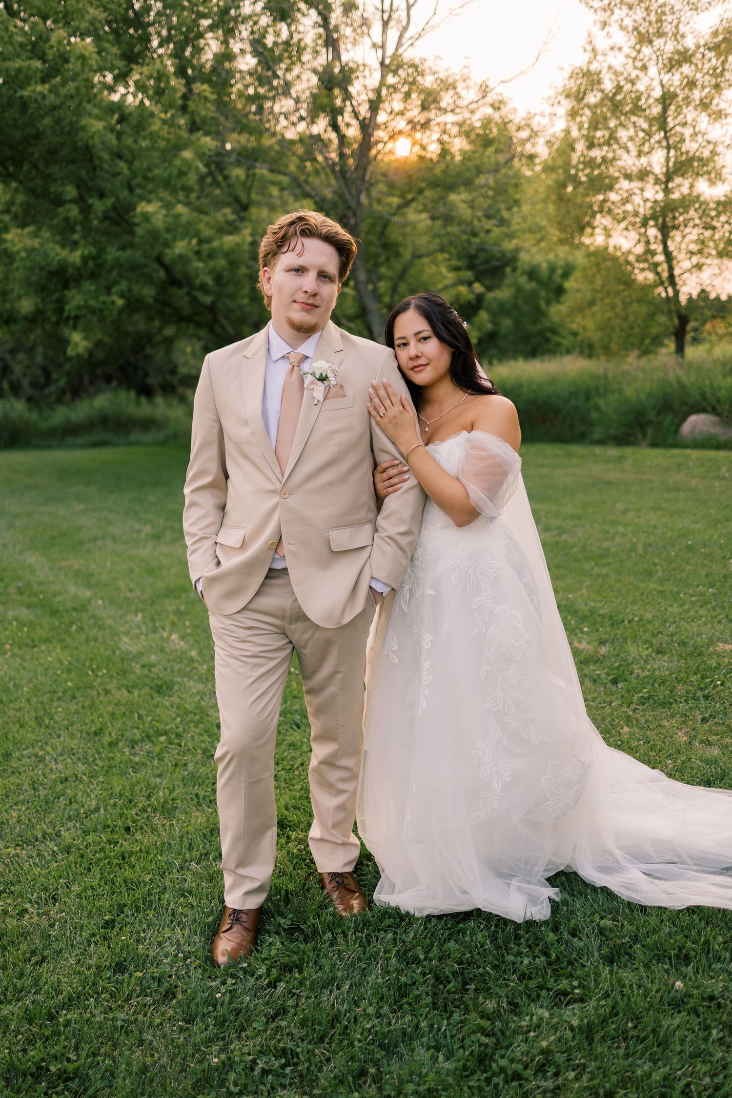 A bride in a white gown and groom in a tan suit pose together on a green lawn at golden hour sunset.