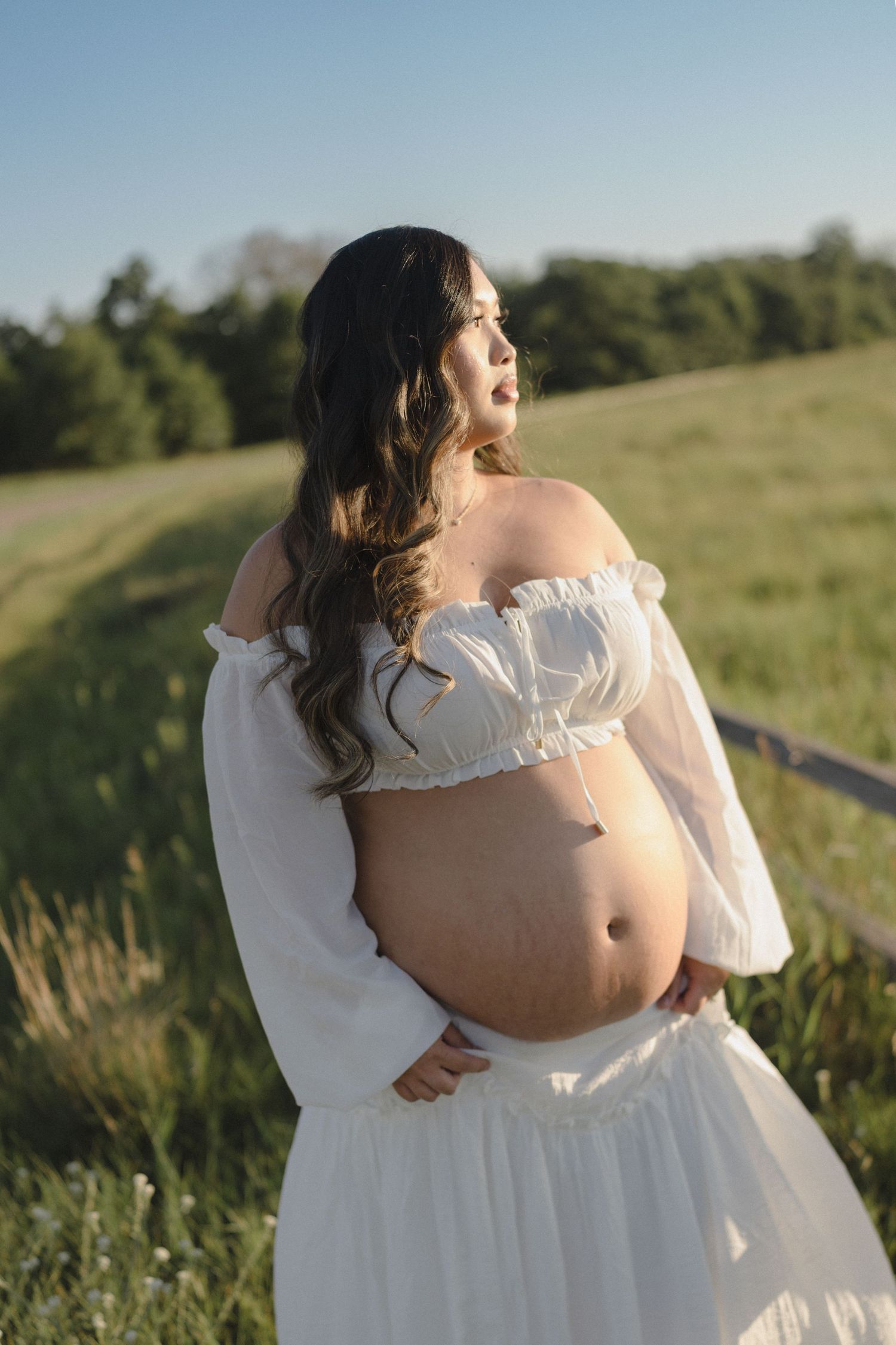 Pregnant woman in white off-shoulder crop top and skirt cradling her baby bump outdoors in a green field.