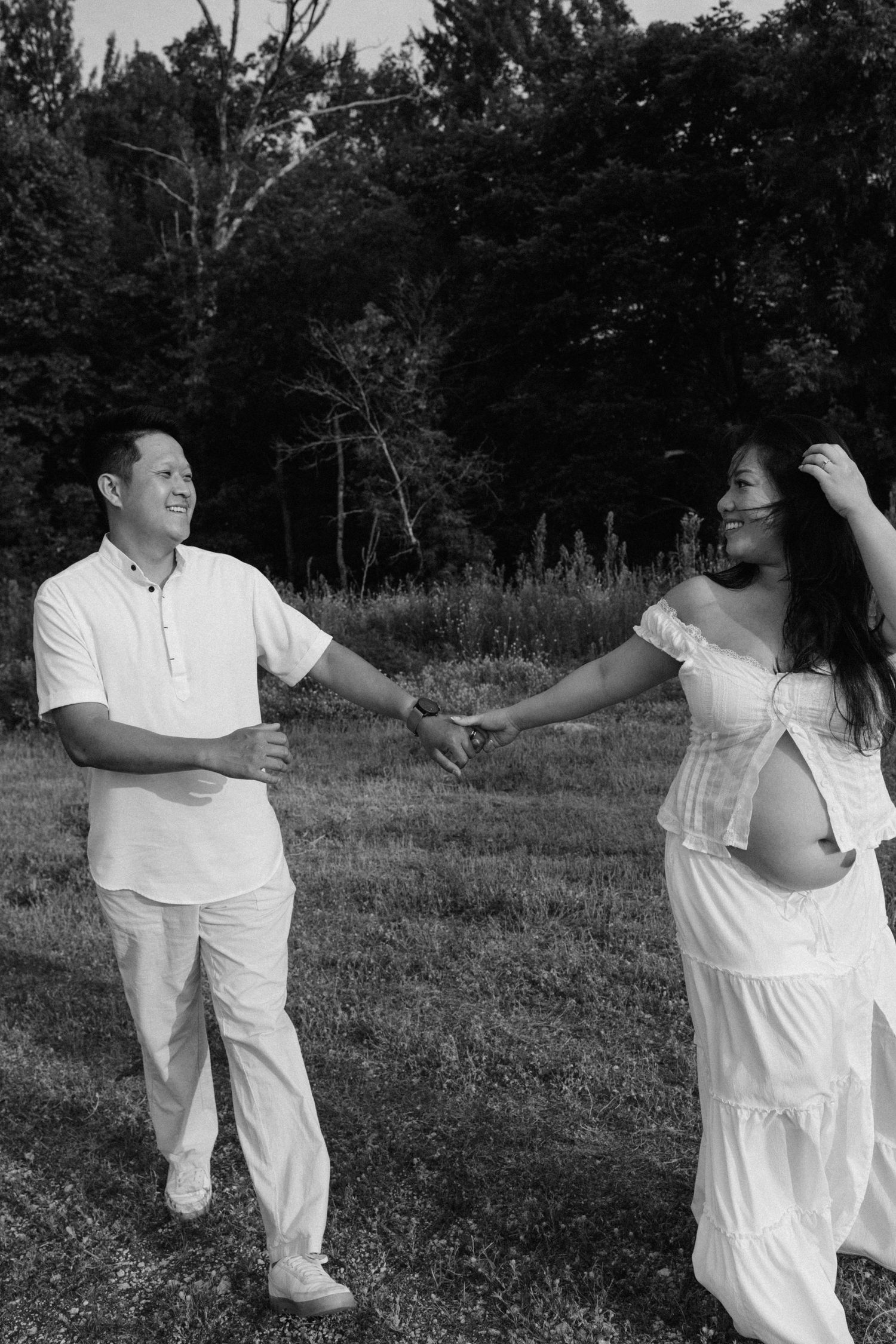 Black and white photo of a smiling couple holding hands in a field, woman in white dress showing pregnant belly.